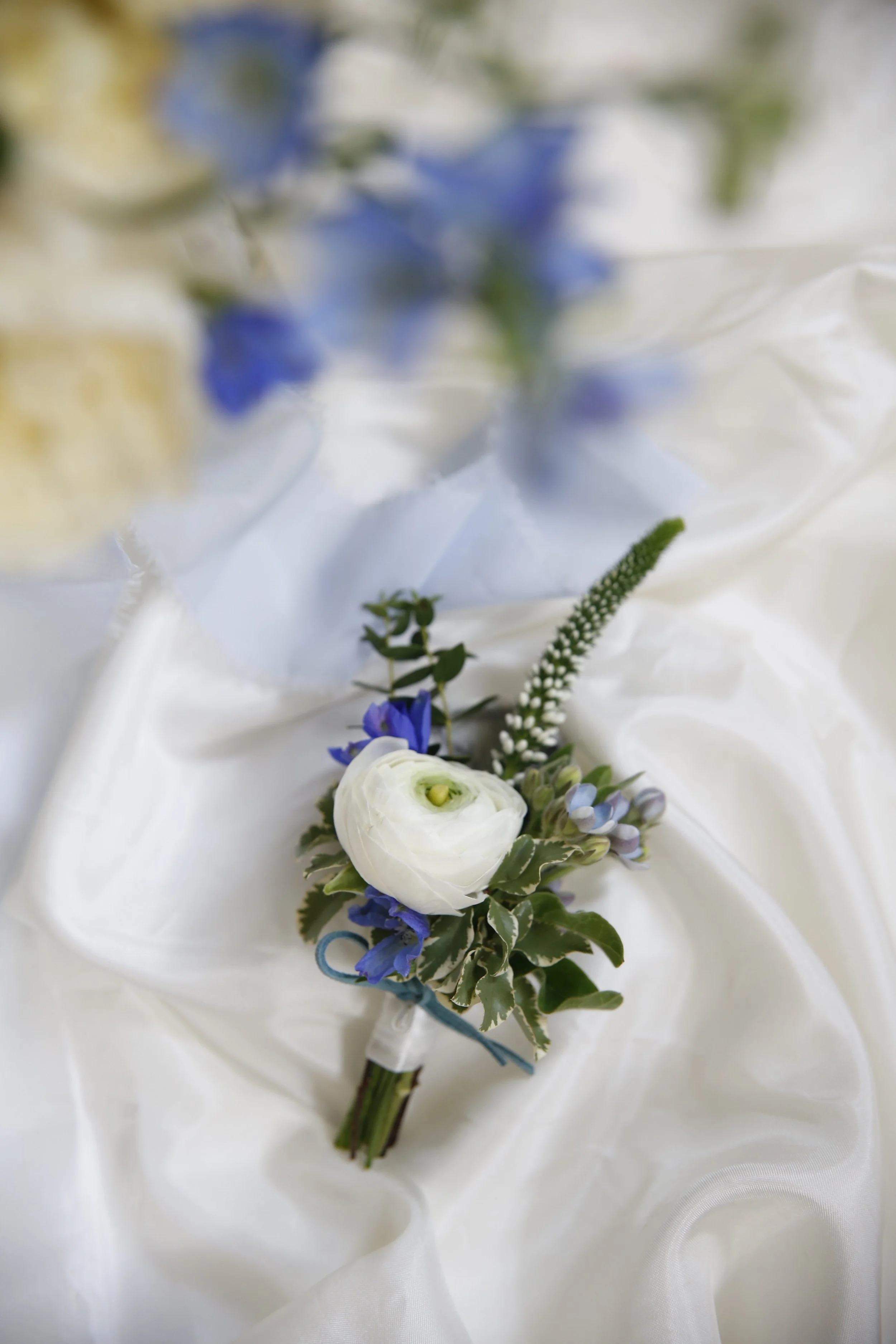 A white and green boutonniere with blue flowers lies on cream-colored satin fabric. The boutonniere includes a white ranunculus, blue delphinium, and greenery, tied with a blue ribbon.