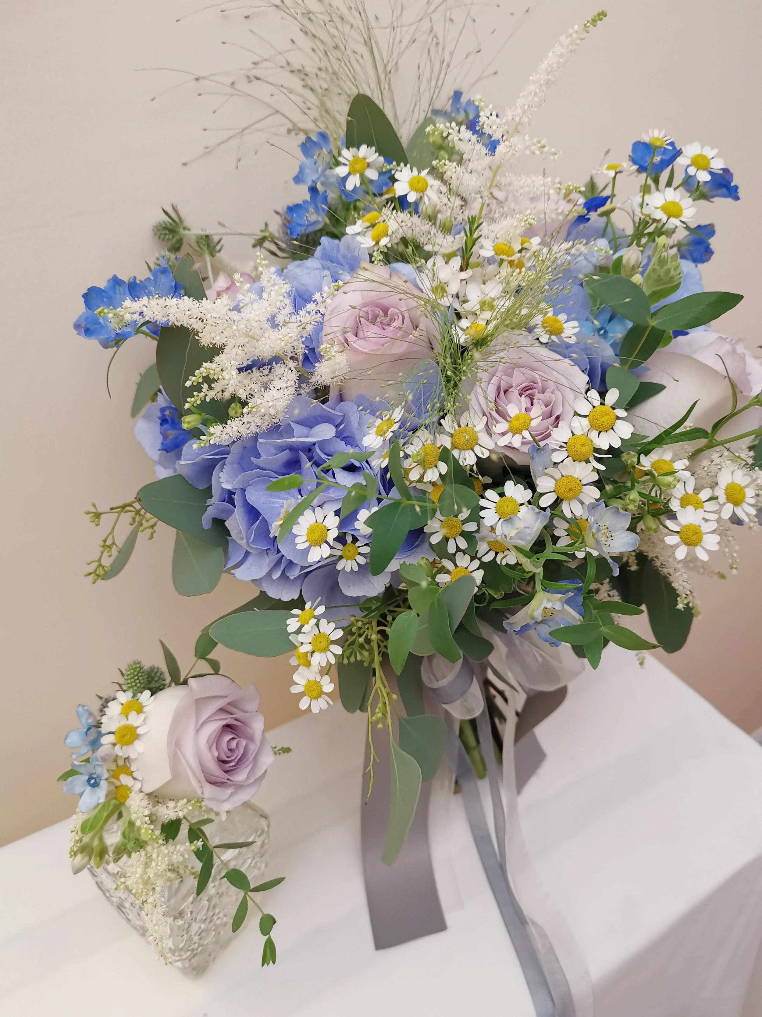 A bouquet of blue, white, and pink flowers with greenery arranged in a large vase, sitting on a white table, with a smaller matching flower arrangement in a glass container nearby.