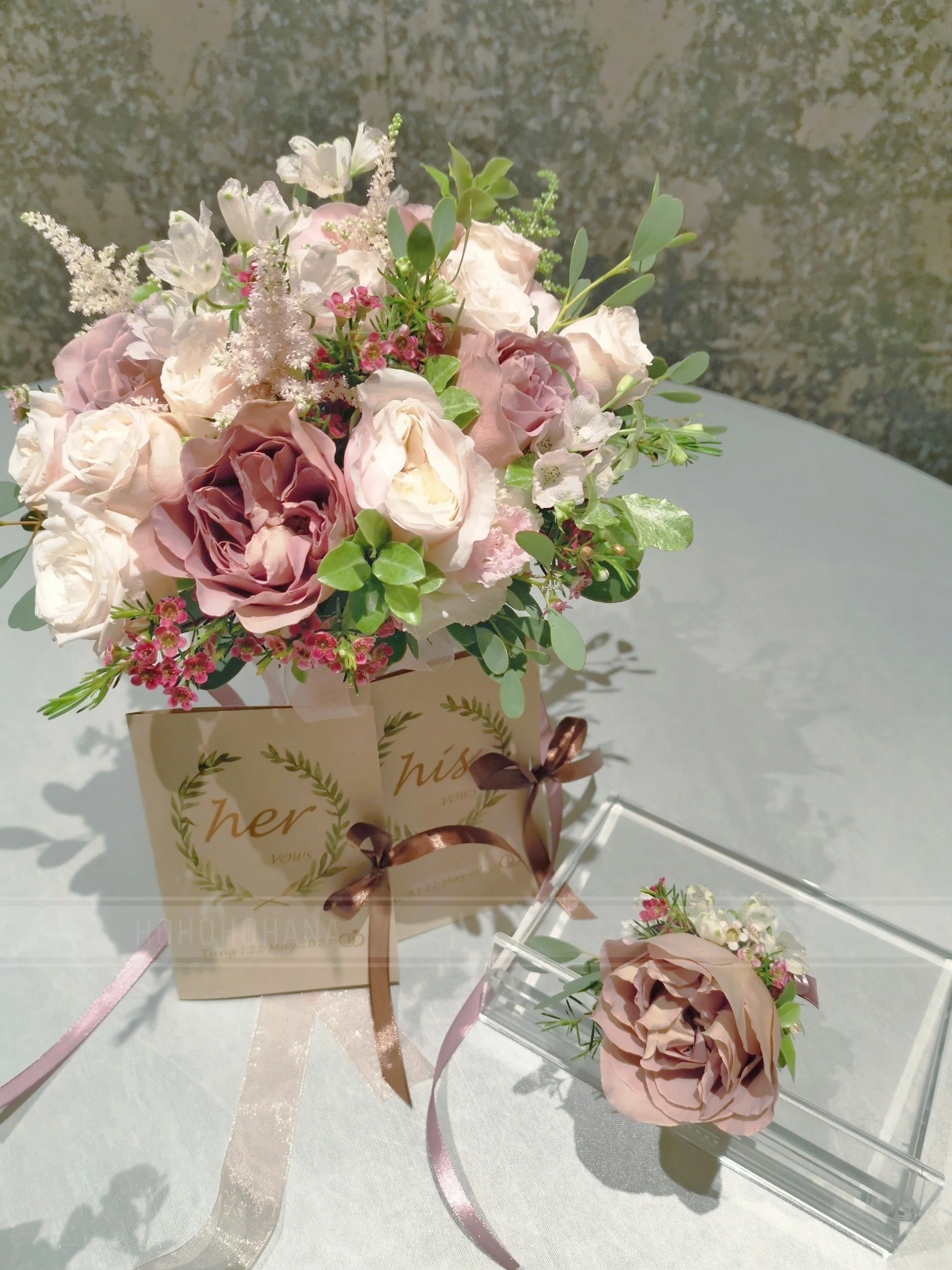 A bouquet of pink, white, and purple flowers with green leaves in a pink box with a ribbon, placed on a white surface, with a single pink flower boutonniere in a small clear holder nearby.