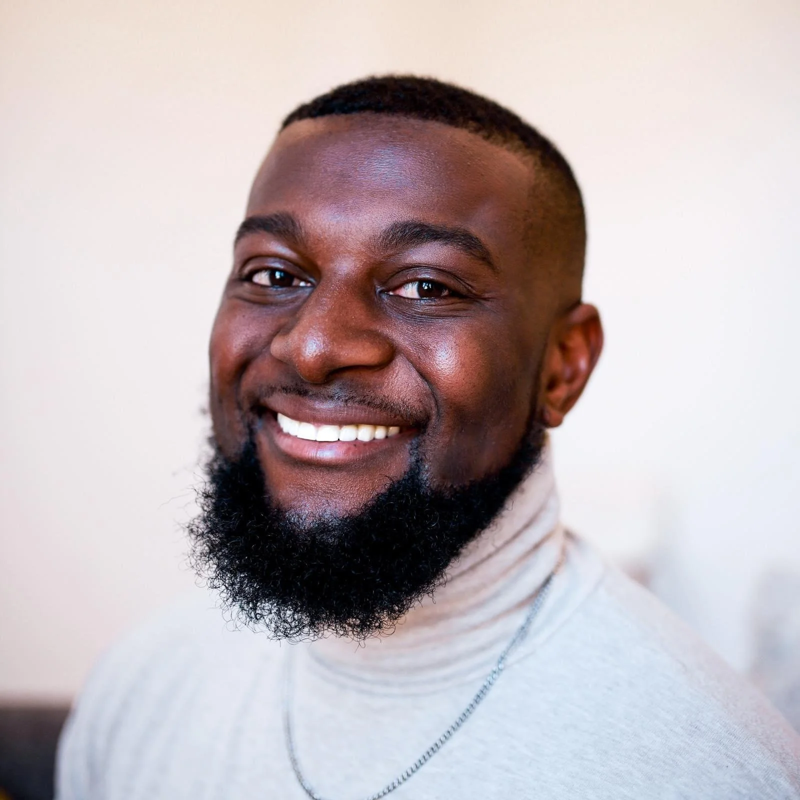Close-up of a smiling African American man with a beard, wearing a light-colored turtleneck and a silver chain, against a plain white background.