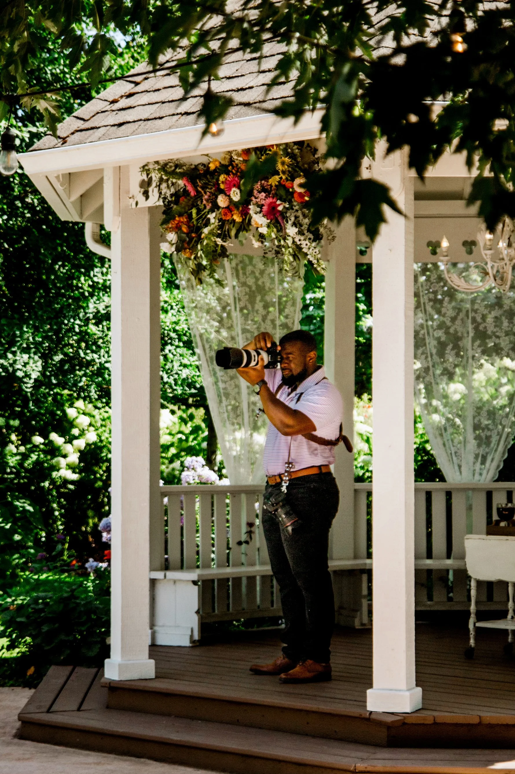 A man is standing on a wooden porch with a white railing, taking a photograph with a professional camera. There are green bushes and flowers in the background, and a floral arrangement hanging above him.