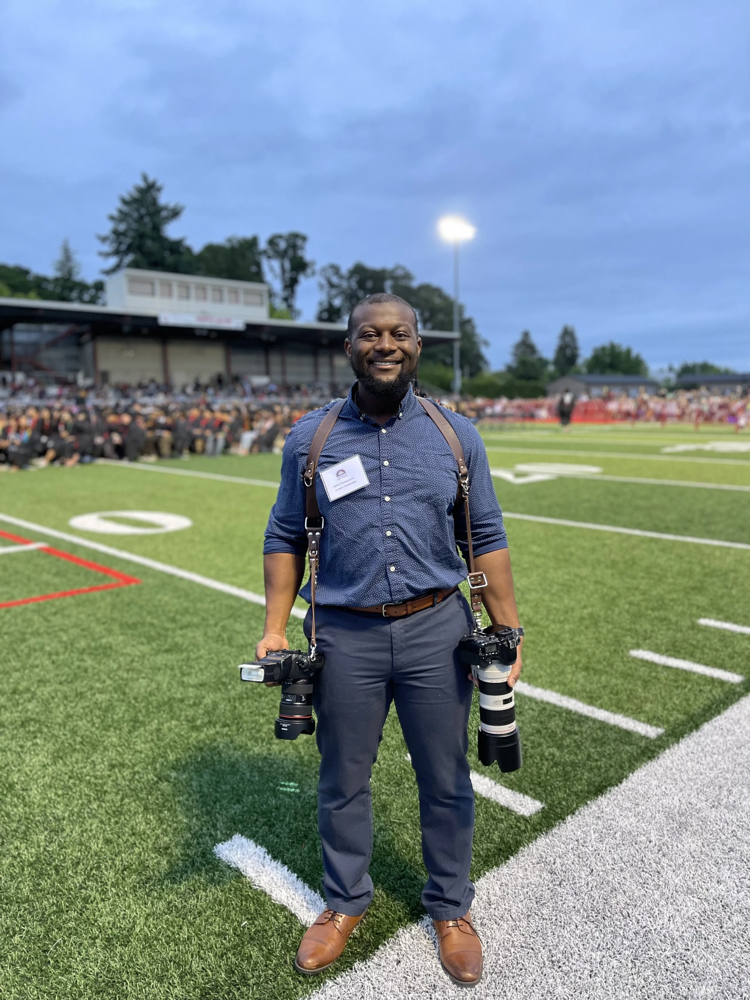 Photographer standing on a sports field with his camera gear, smiling, during evening with stadium lights and a crowd in the background.