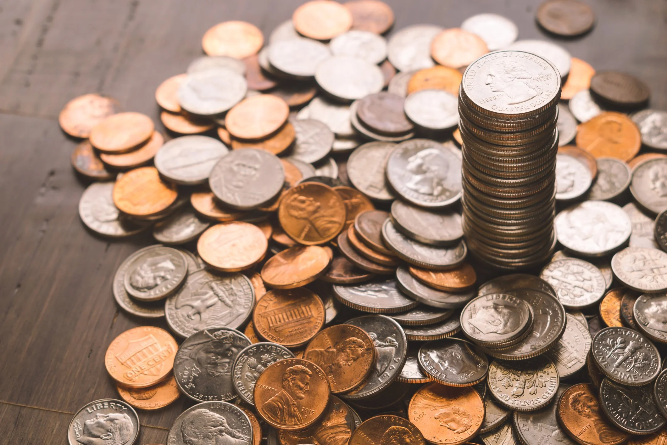 A pile of various U.S. coins, including pennies, nickels, and quarters, with a stack of quarters positioned vertically on top, on a wooden surface.