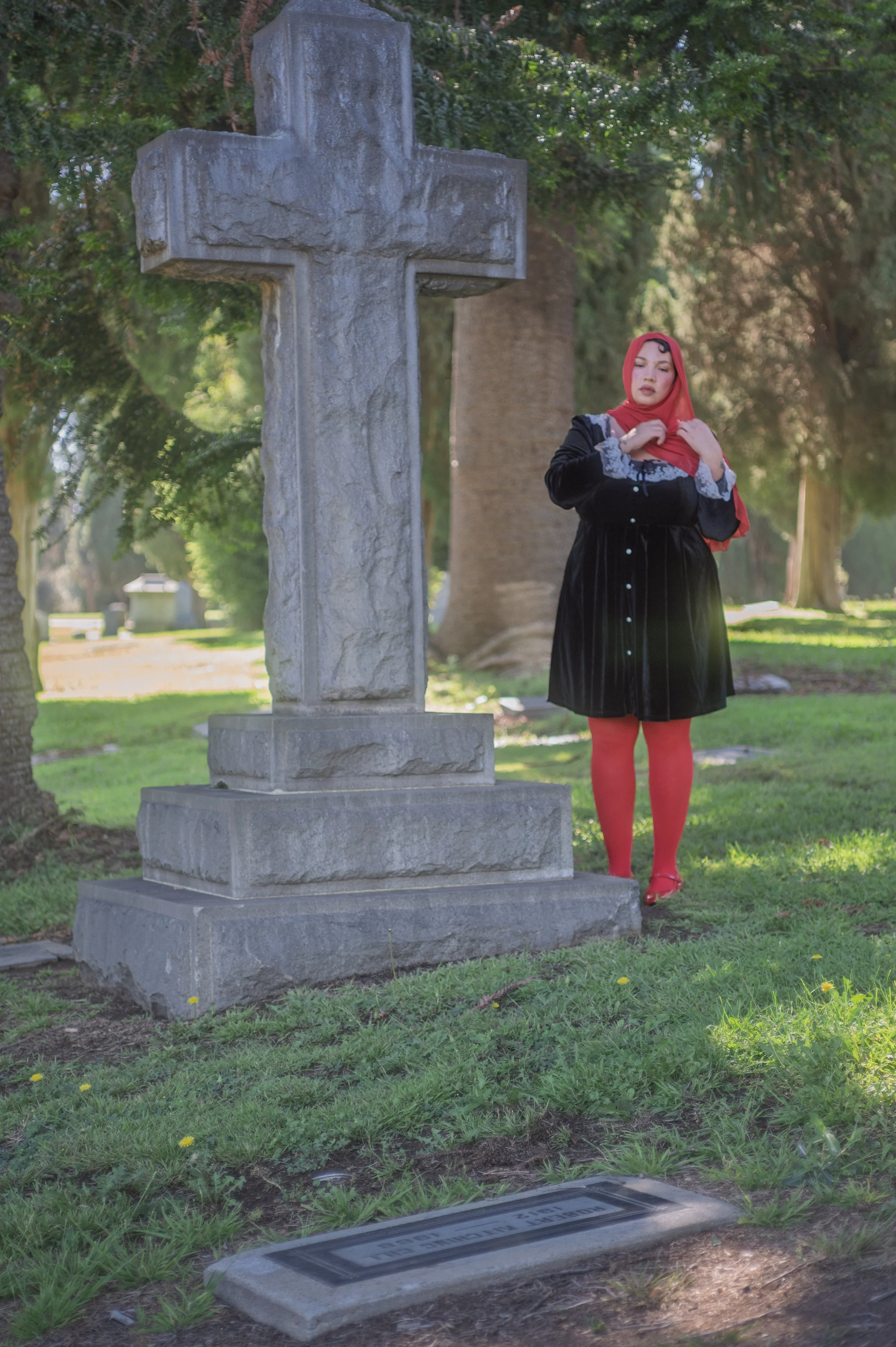 A young woman with medium skin tone standing outdoors near a large stone cross monument and a grave marker with the inscription 'FAMILY OF'. She is wearing a red headscarf, black dress with lace details, red tights, and red shoes. She appears to be in a cemetery during daytime, surrounded by trees and greenery.