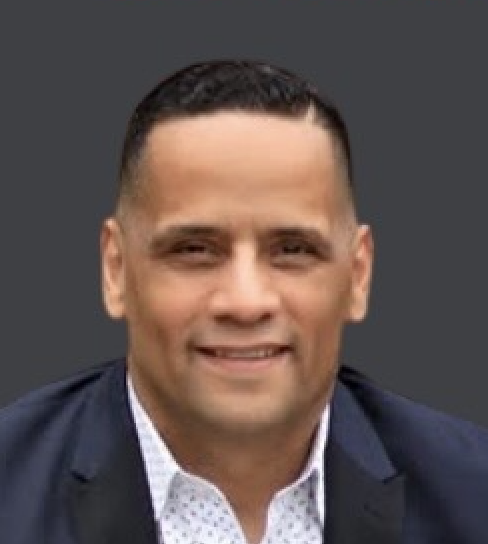 Headshot of a man with short dark hair wearing a dark blazer and a white collared shirt against a gray background.