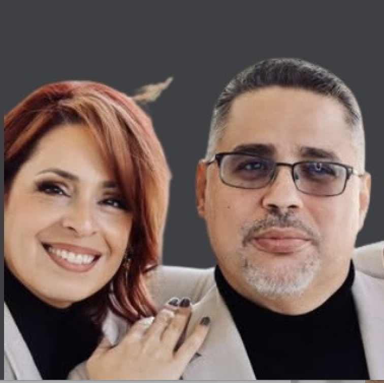 A smiling woman with red hair and a man with glasses and gray hair, both dressed in light-colored suits, posing closely together against a dark background.