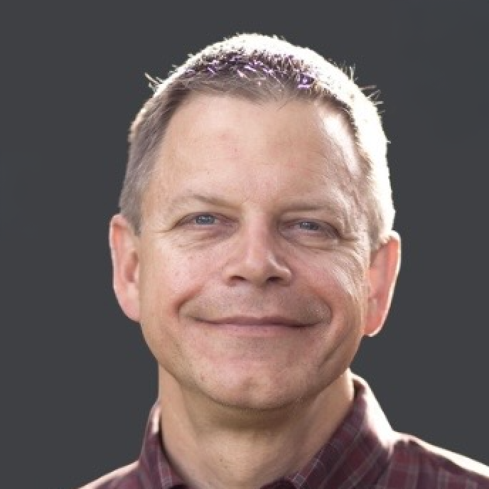 Portrait of a middle-aged man with short hair, smiling, wearing a maroon plaid shirt, against a dark background.