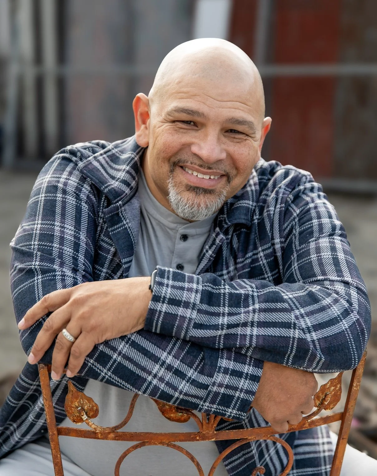 A smiling bald man with a goatee, wearing a plaid shirt over a light grey T-shirt, sitting outdoors leaning on a rusty metal chair.