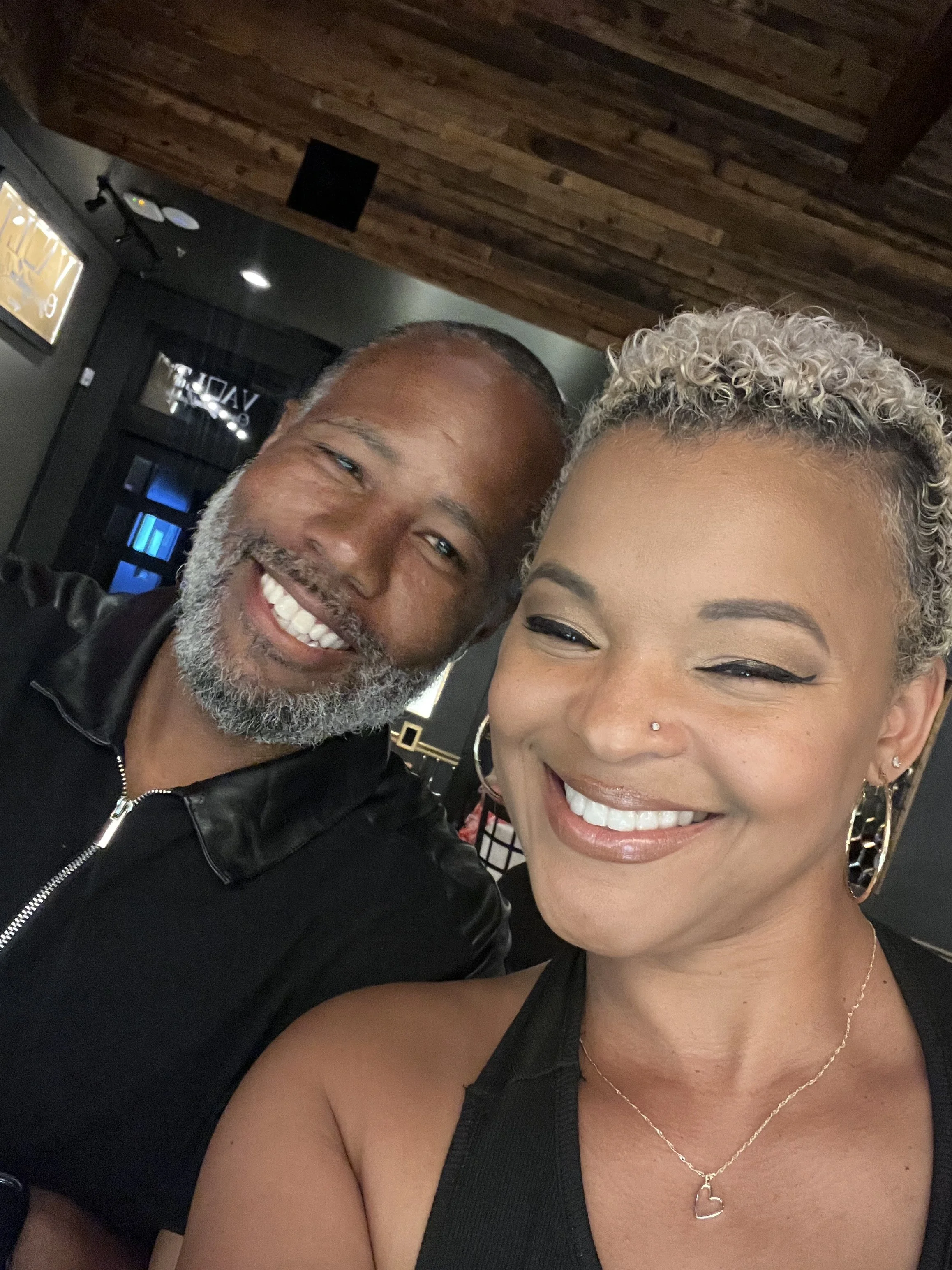 A smiling man with a beard and a woman with short, curly blonde hair, both excitedly posing for a selfie in a dimly lit indoor setting with a wooden ceiling.