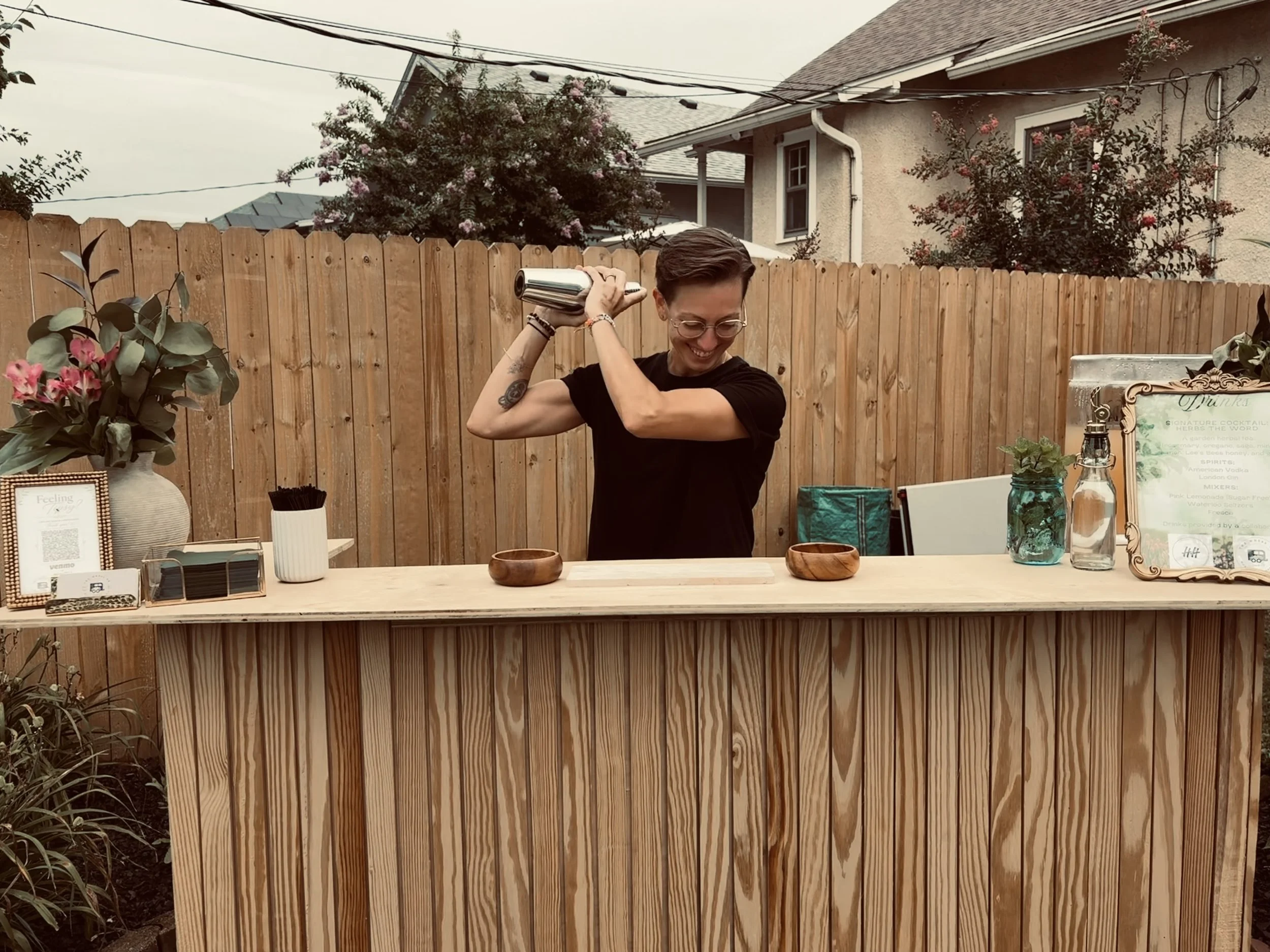 A smiling person with short hair, glasses, and tattoos on their arm, mixing drinks at an outdoor bar with a wooden counter, surrounded by plants, decorative items, and a menu sign, with a house and trees in the background.