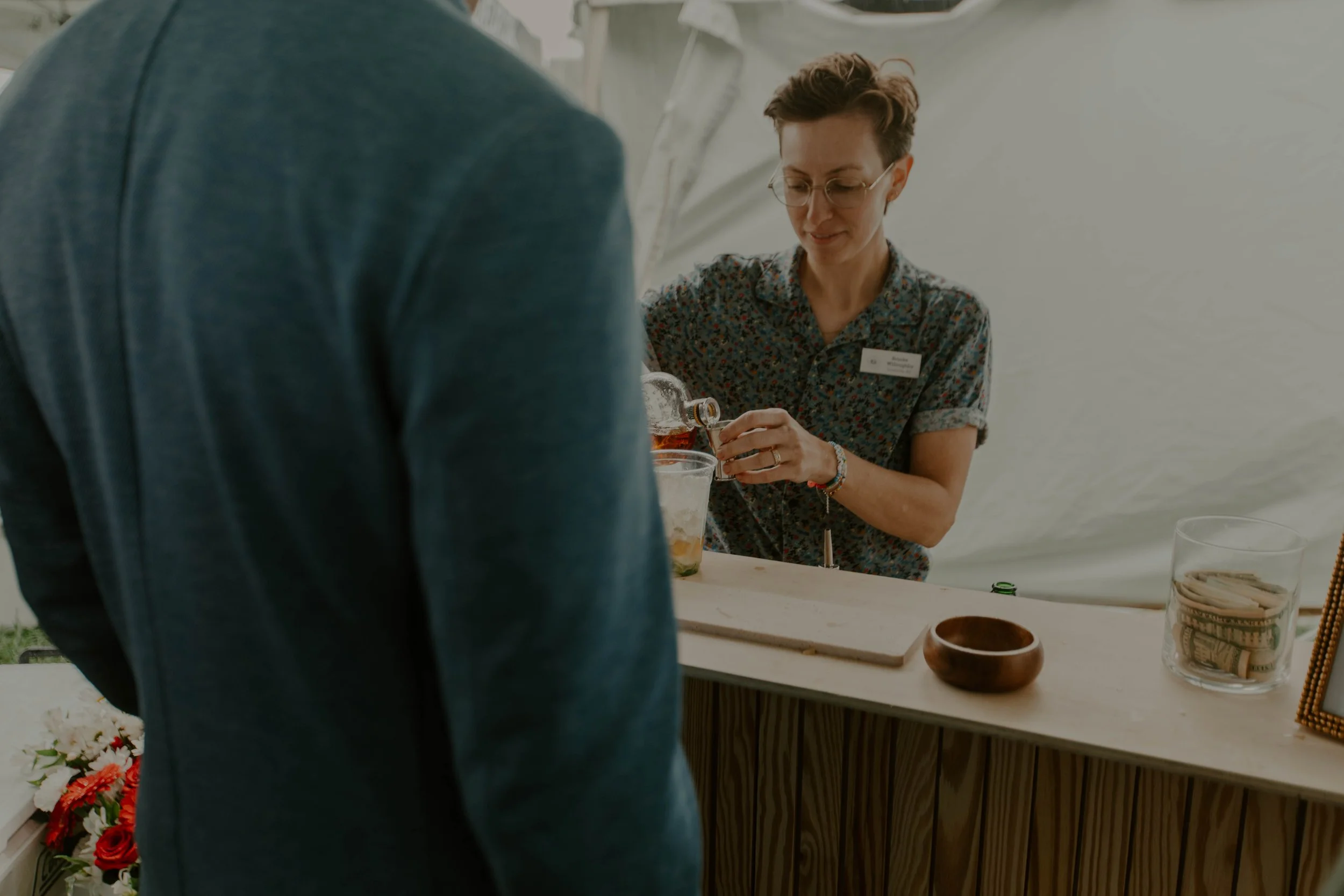 A woman with short hair and glasses, wearing a floral shirt, pouring a drink into a glass at a table. A man in a teal shirt stands in front of her, with flowers visible to the left and various items on the table, including a basket of rolled items, a glass jar with a straw, and a wooden bowl.