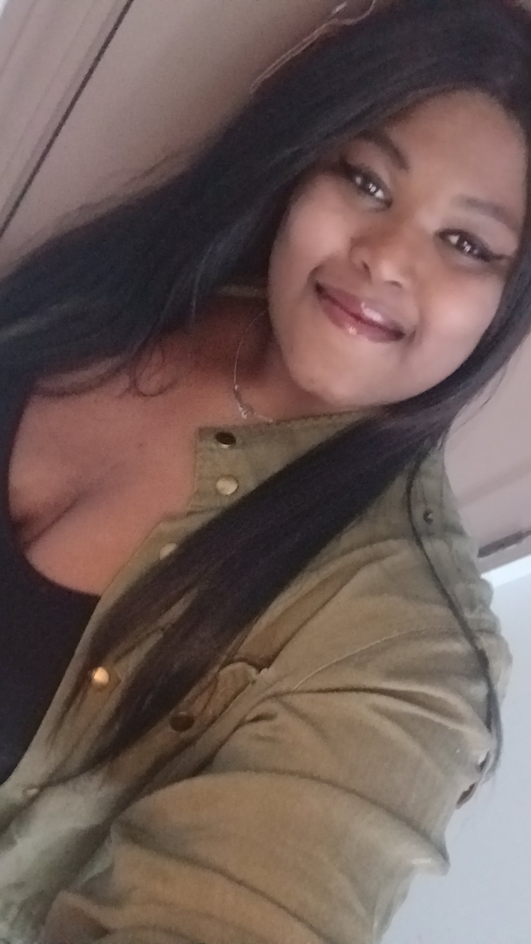 A young woman with long black hair smiling at the camera, wearing a black top and a tan jacket with metallic buttons.