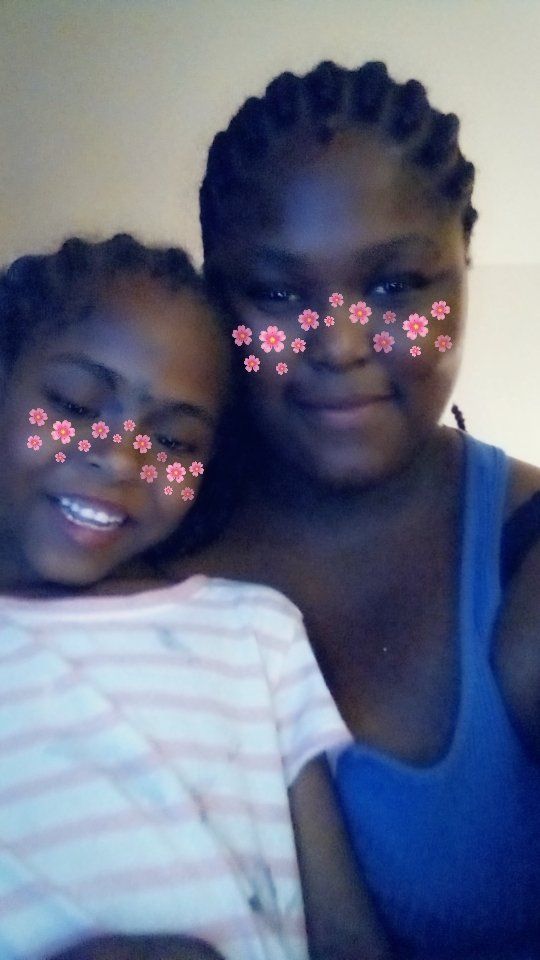 Two young girls smiling, with pink flower filters on their faces, close together for a photo. The girl on the left is wearing a striped shirt and the girl on the right is in a blue tank top.