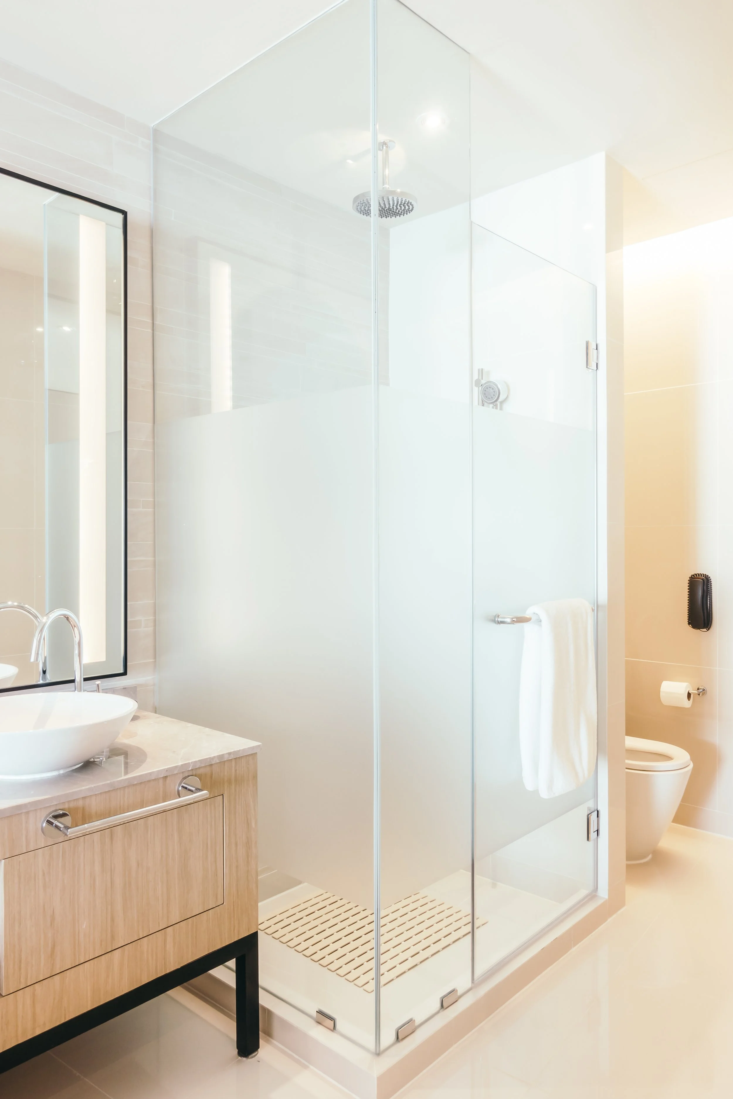 Modern bathroom with a glass shower enclosure, a white sink on a wooden vanity, a mirror, and a toilet with a towel and toilet paper.