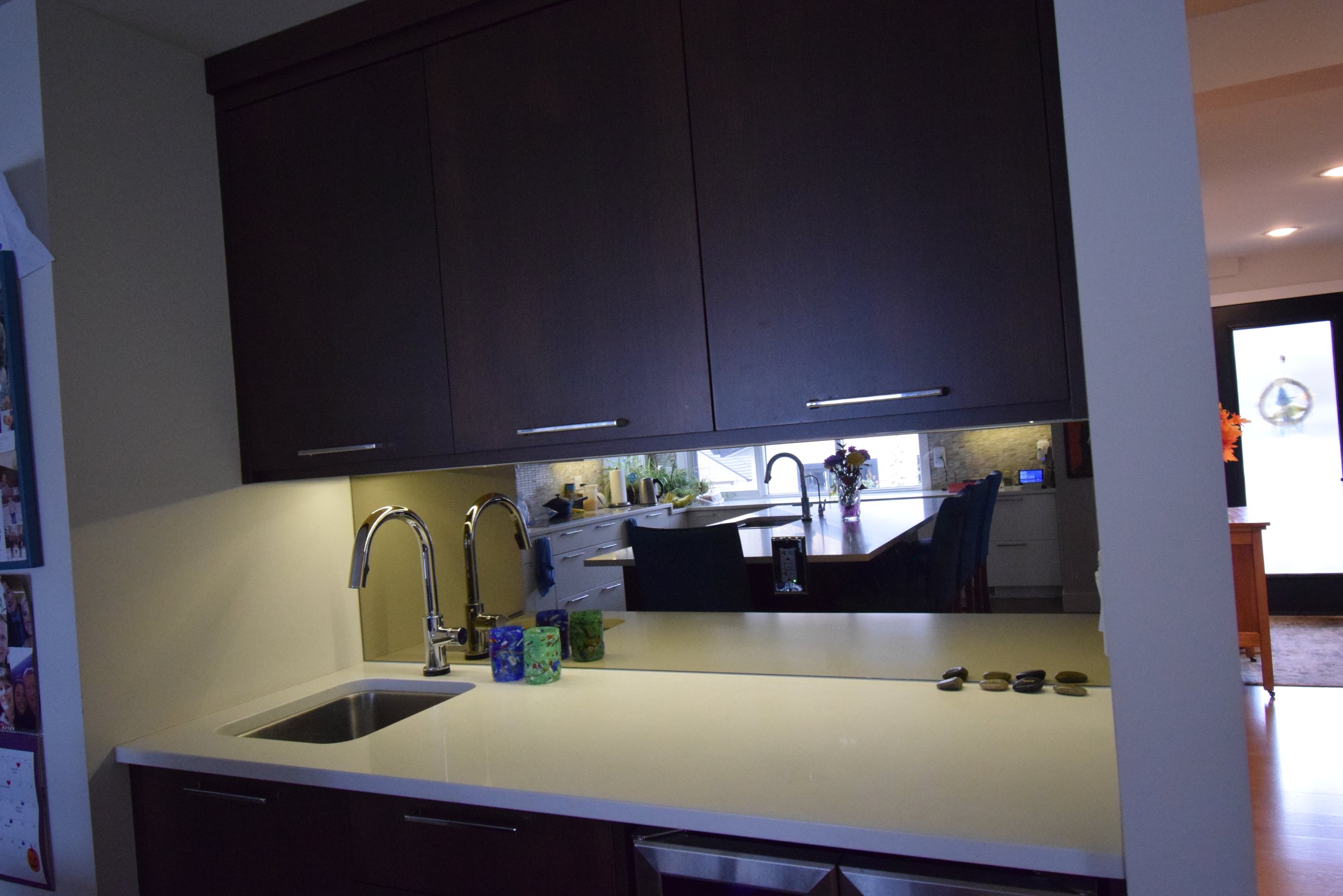 A kitchen with a white countertop, two silver faucets, a small sink, and colorful mugs, with a view into the dining area and living room in the background.