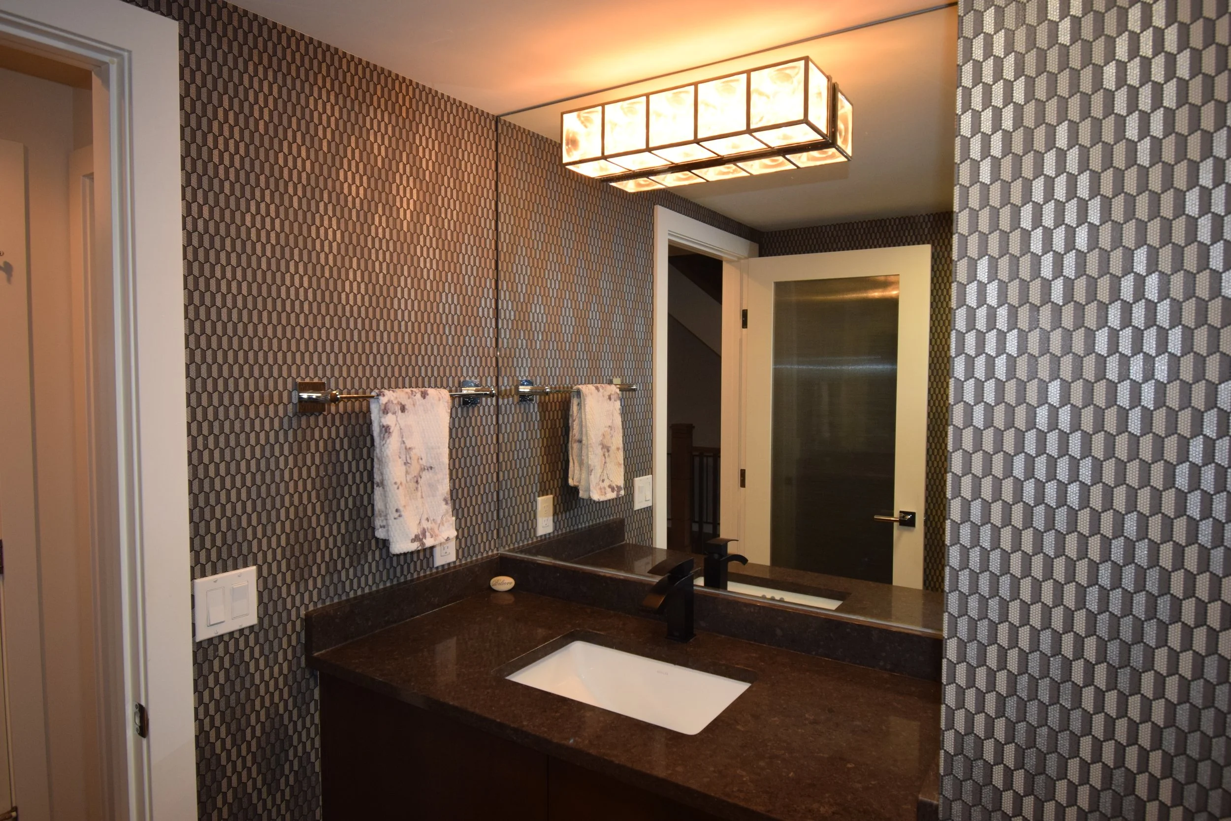 Bathroom with hexagonal tile wallpaper, dark countertop, white sink, black faucet, towel rail with two towels, large mirror, ceiling light fixture, door with frosted glass, and light switch.