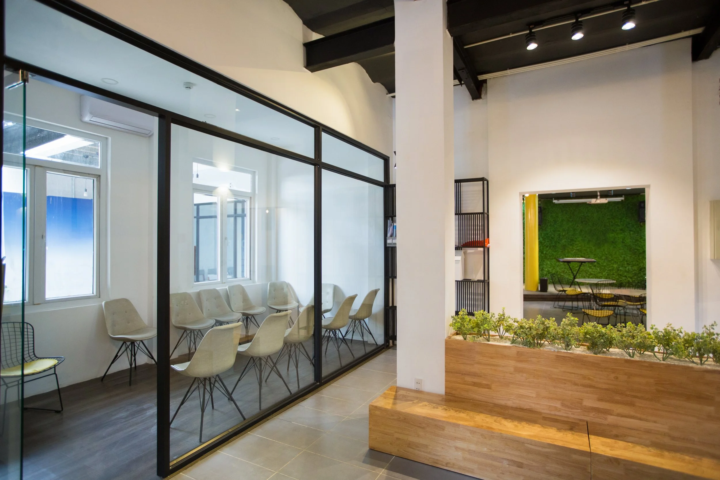 Modern office interior with glass-walled conference room containing white chairs around a table, a window with natural light, a wooden planter with small green plants, and a separate room with a green grass wall, yellow pillars, and tables with yello