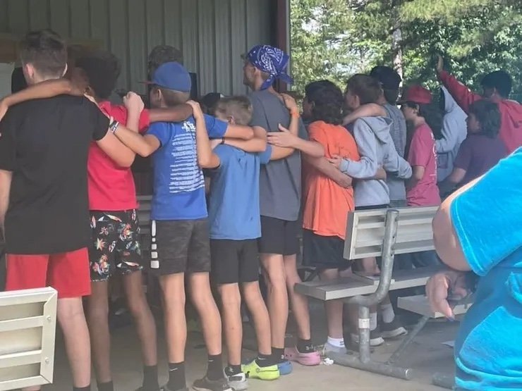 A group of children standing in a line, arms around each other's shoulders, at a summer camp or outdoor event.