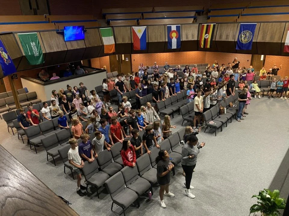 Children and adults participating in a church service or prayer session in a large auditorium with flags hanging from the balcony.
