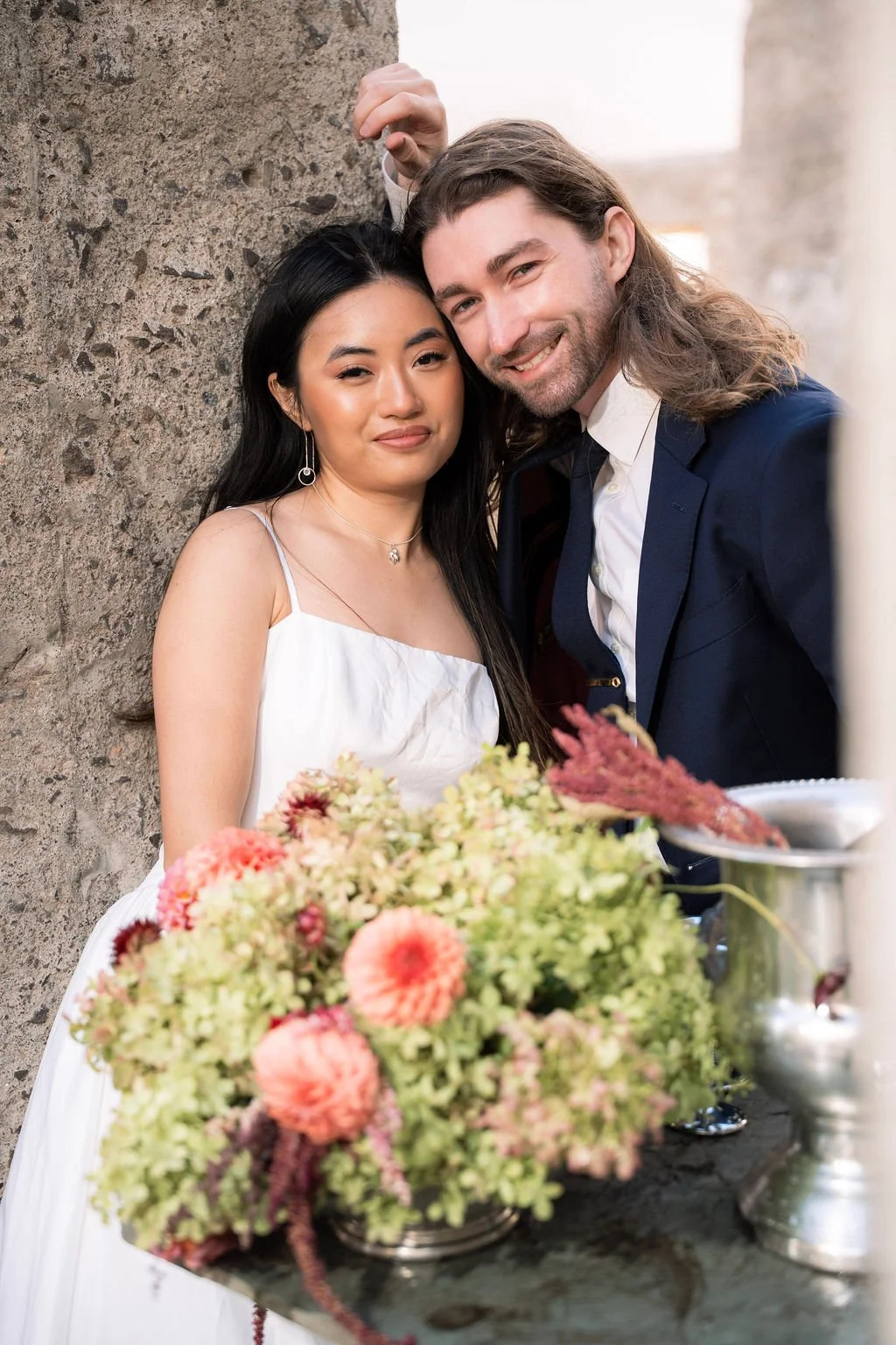 A couple standing close together outdoors, with a floral arrangement and a silver punch bowl in front of them, near a textured stone wall.