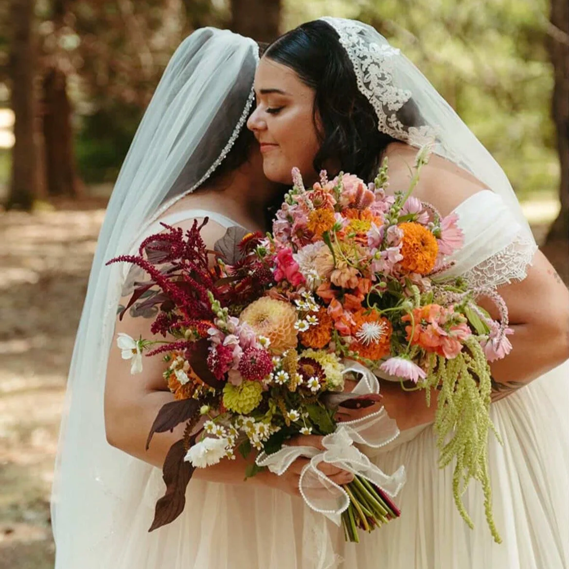 Two women in wedding dresses sharing a hug, with one holding a colorful bouquet of flowers, outdoors in a wooded area.