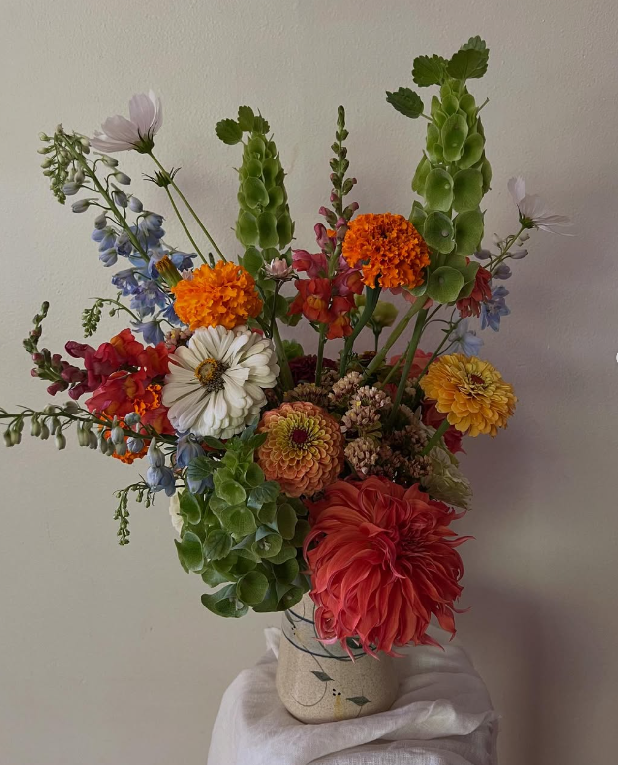A colorful bouquet of various flowers in a decorative vase on a white cloth-covered surface against a plain wall background.