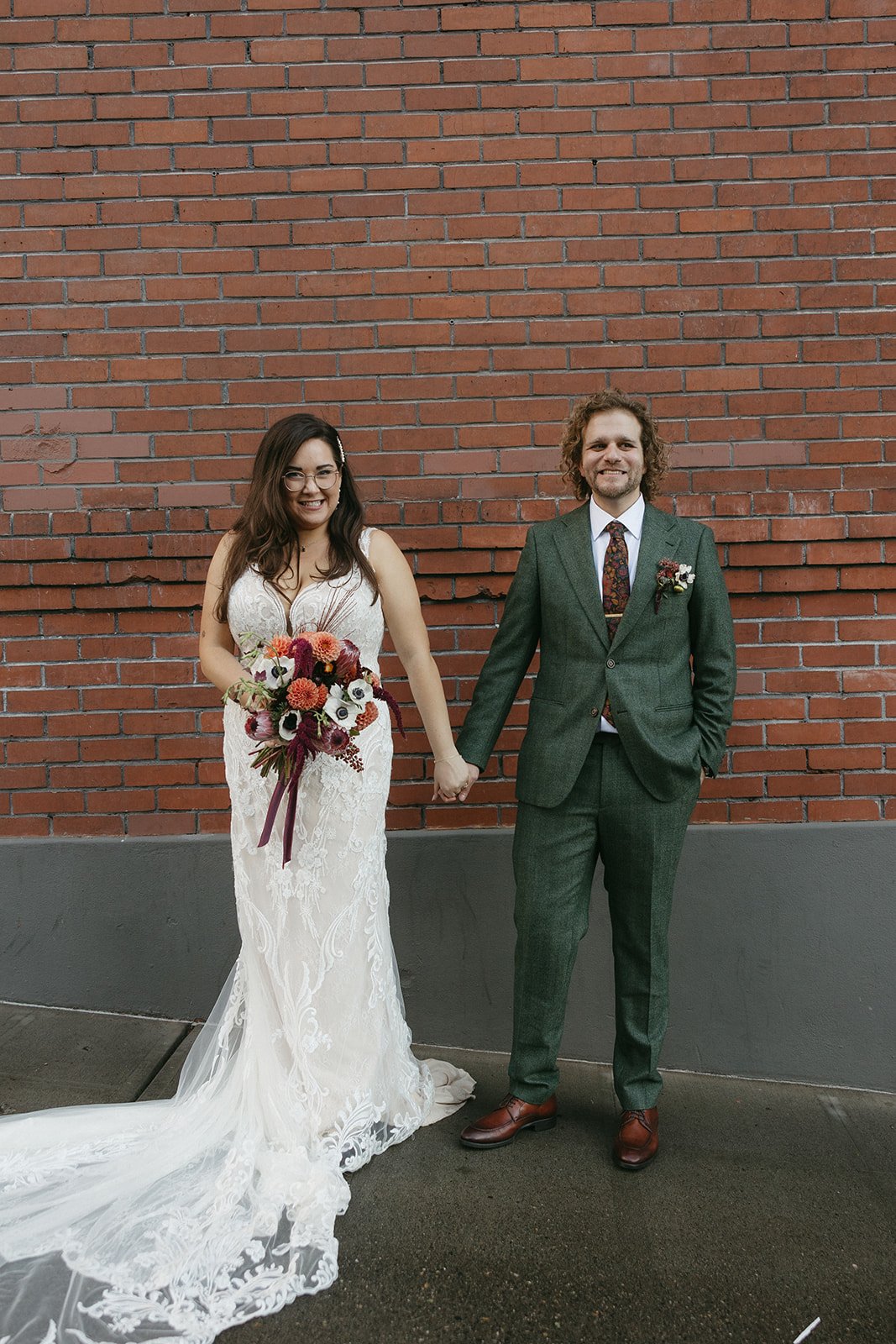 A newly married couple standing against a brick wall, holding hands and smiling. The bride is in a white lace wedding dress and holding a bouquet of vibrant flowers. The groom is in a green suit with a floral-patterned tie and a boutonniere.