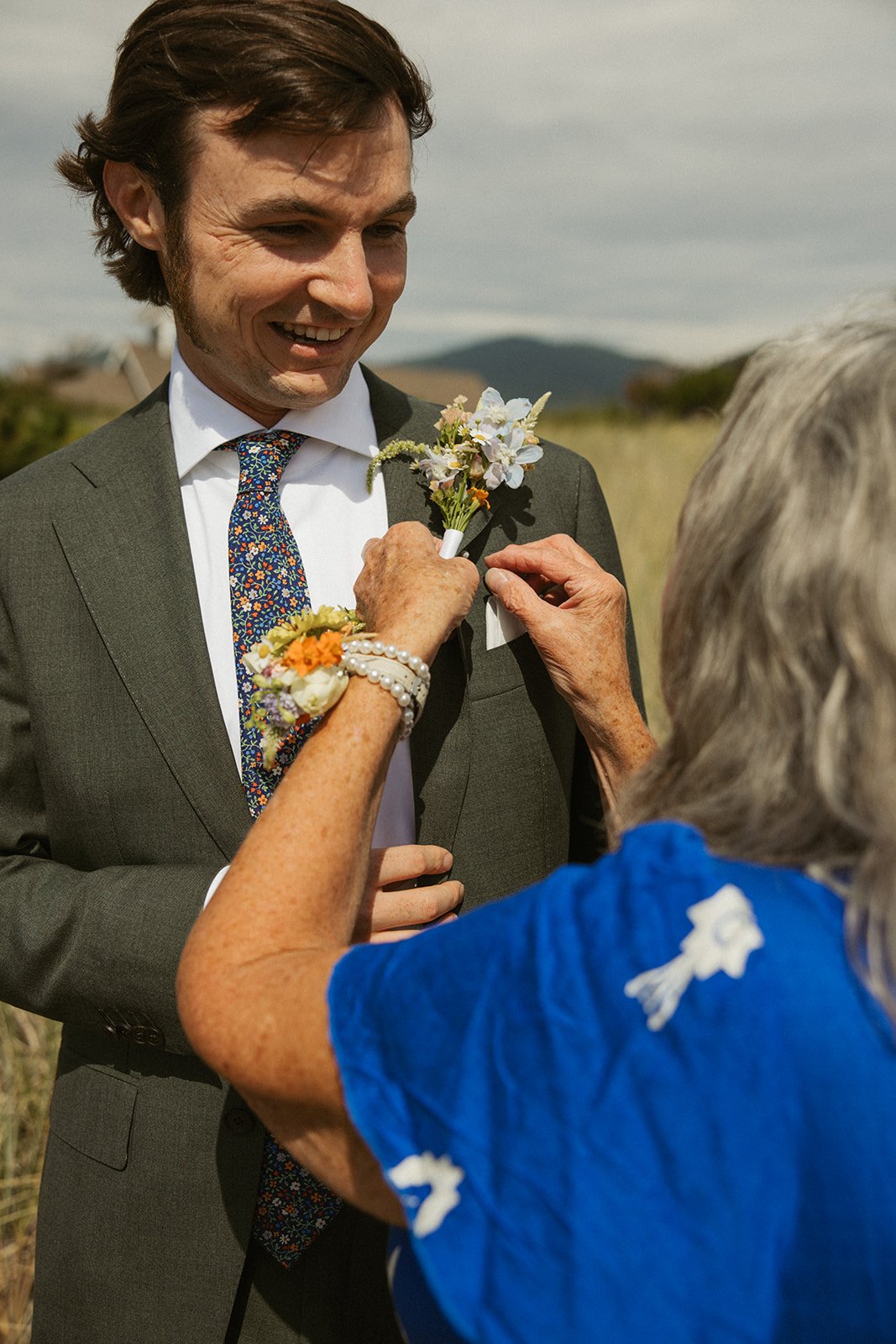 An older woman pins a boutonniere on a young man wearing a suit and tie outdoors, likely at a wedding or special event.