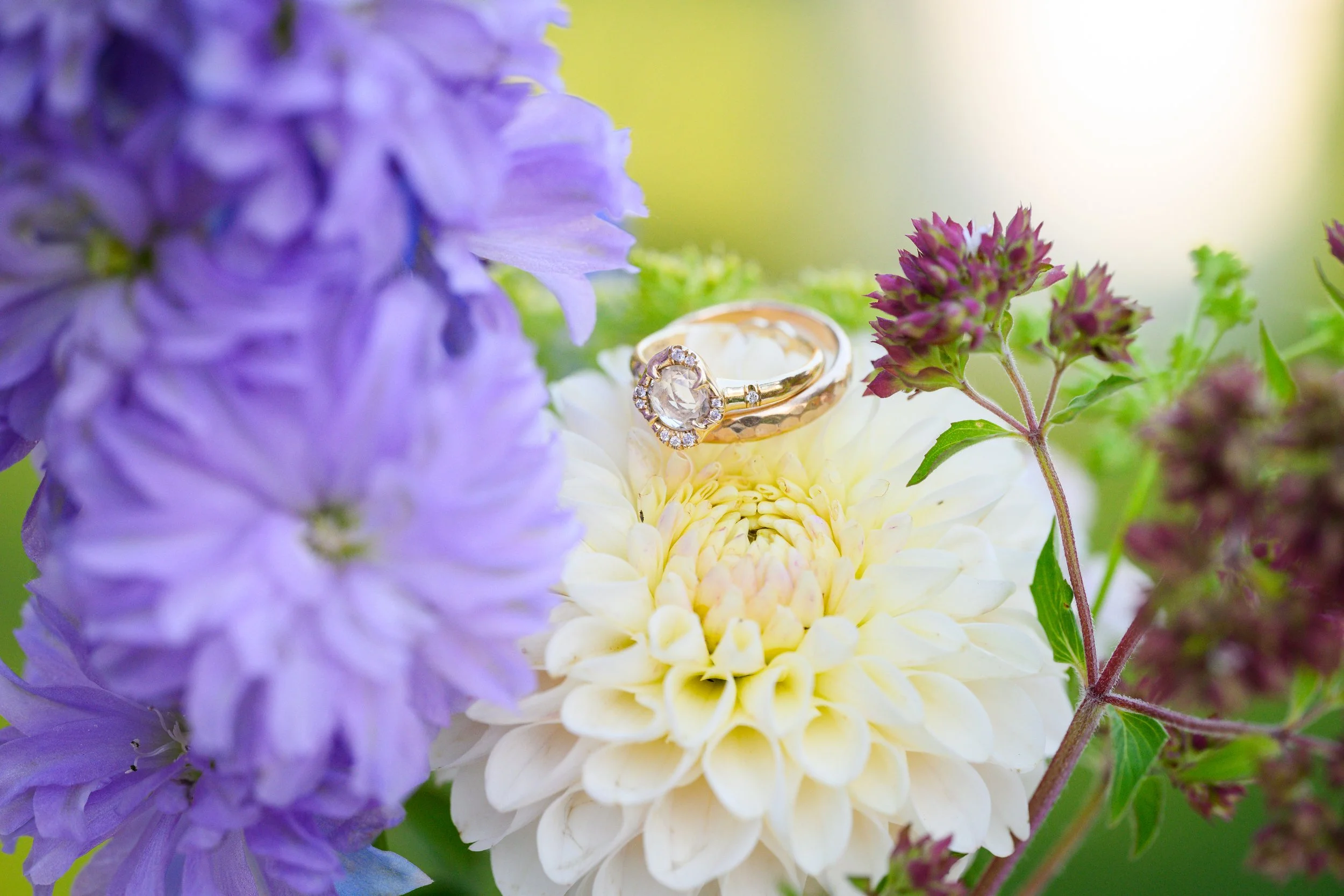 A close-up of a gold engagement ring with a large central diamond and smaller surrounding diamonds, resting on a white flower among purple and green flowers.