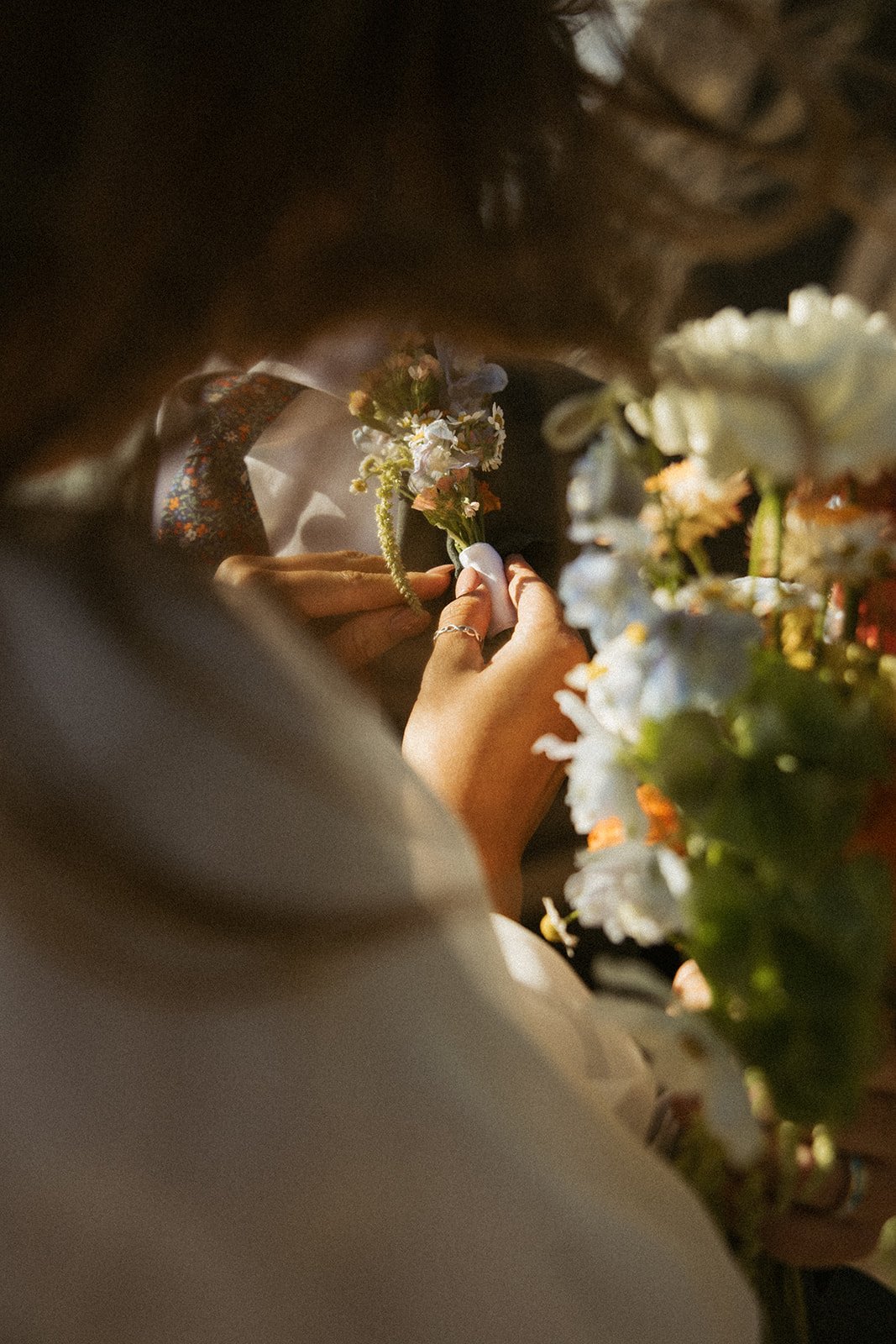 Close-up of a person arranging a small bouquet of flowers, with other flowers visible in the foreground, and a person wearing a suit and floral tie in the background.