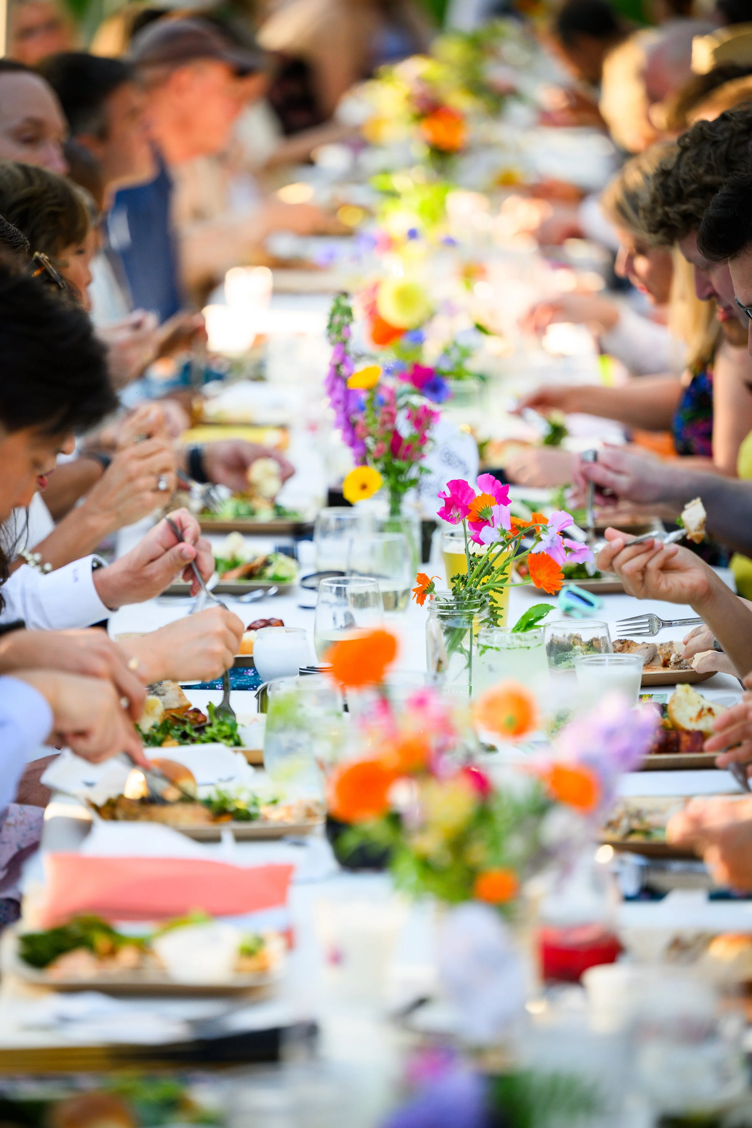 People dining outdoors at a long table decorated with colorful flower arrangements.