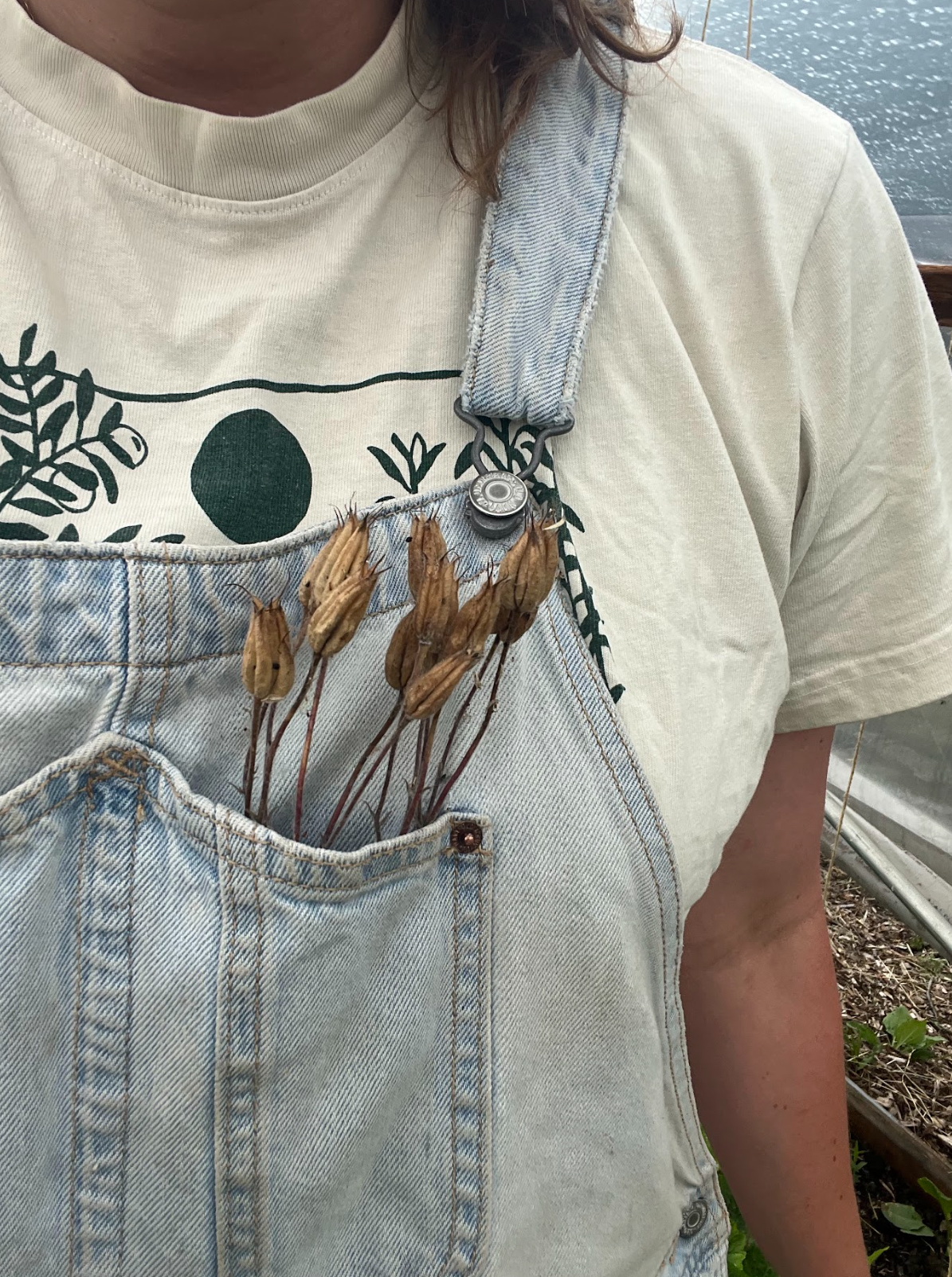 Close-up of a person wearing a white t-shirt with a graphic design and light blue denim overalls. The overalls have a chest pocket filled with dried seed pods. The person is outdoors near water.