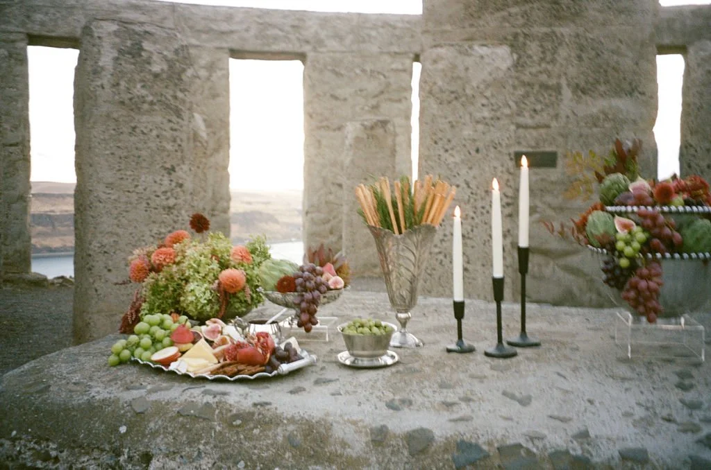A stone table set with floral arrangements, grapes, and cheeses, with four white candles in black holders and a vase of breadsticks, outdoors with stone ruins and a view of water in the background.