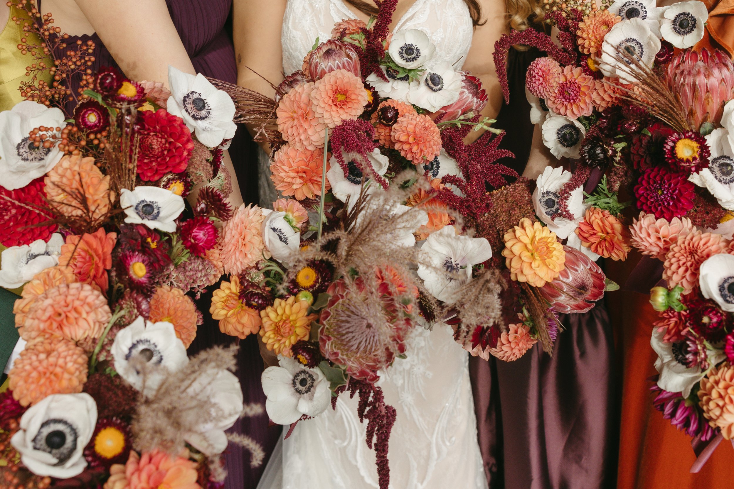 Close-up of a bride holding a large, colorful floral bouquet with peach, red, white, and burgundy flowers, surrounded by bridesmaids with similar bouquets.