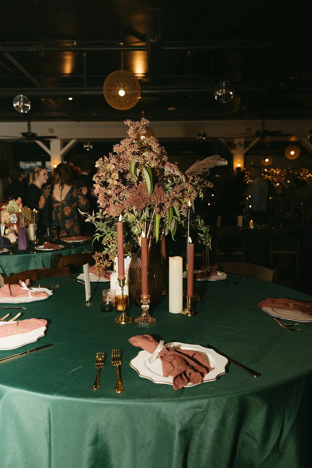 A decorated round table with a green tablecloth, dressed with pink napkins and gold cutlery, featuring a centerpiece of pink and purple flowers in a dark vase, surrounded by candles and glasses, set for a formal event in a dimly lit venue with people