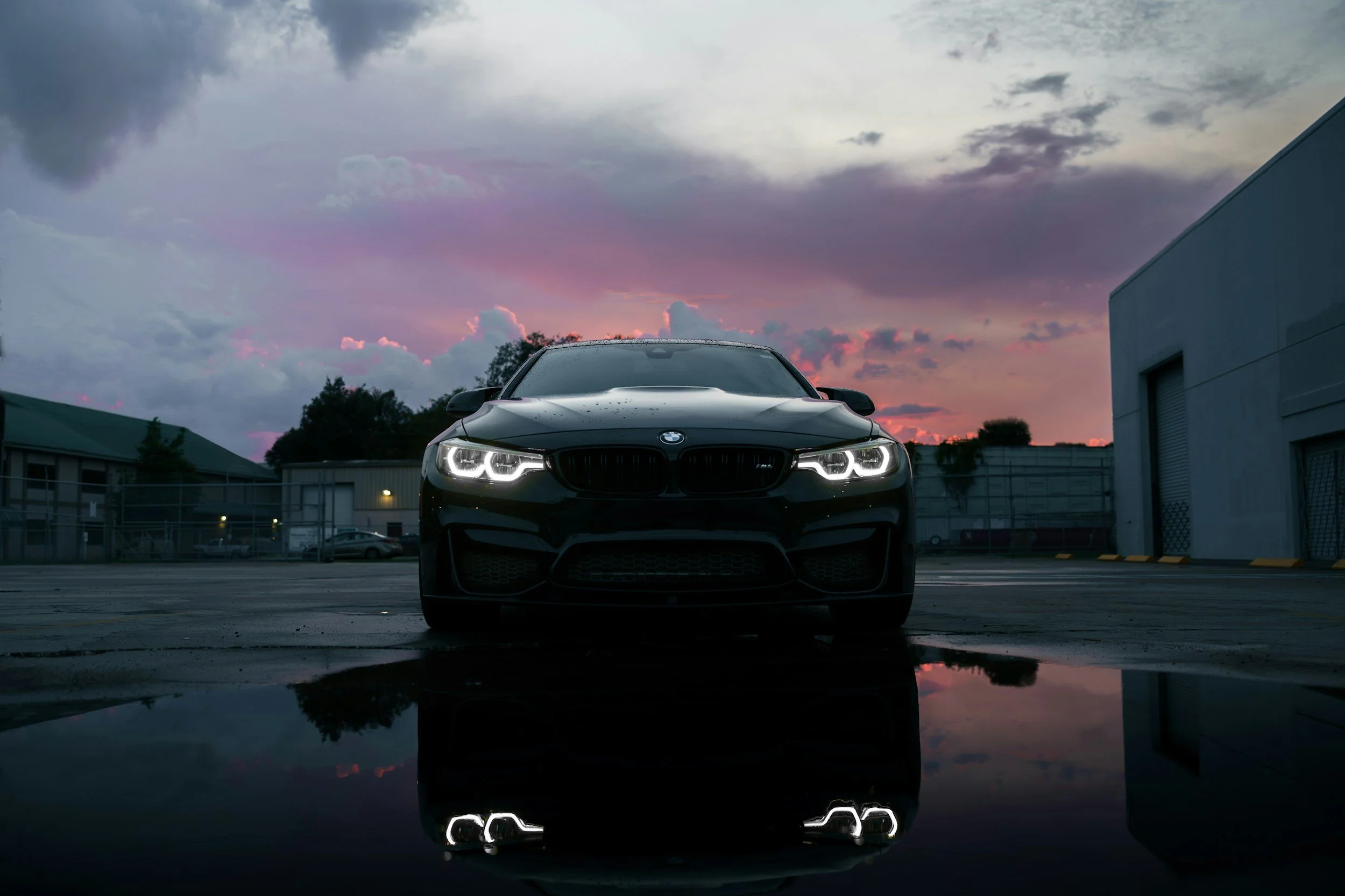 Black sports car parked in an industrial area at sunset, with its headlights on and reflection visible on a wet surface in front.