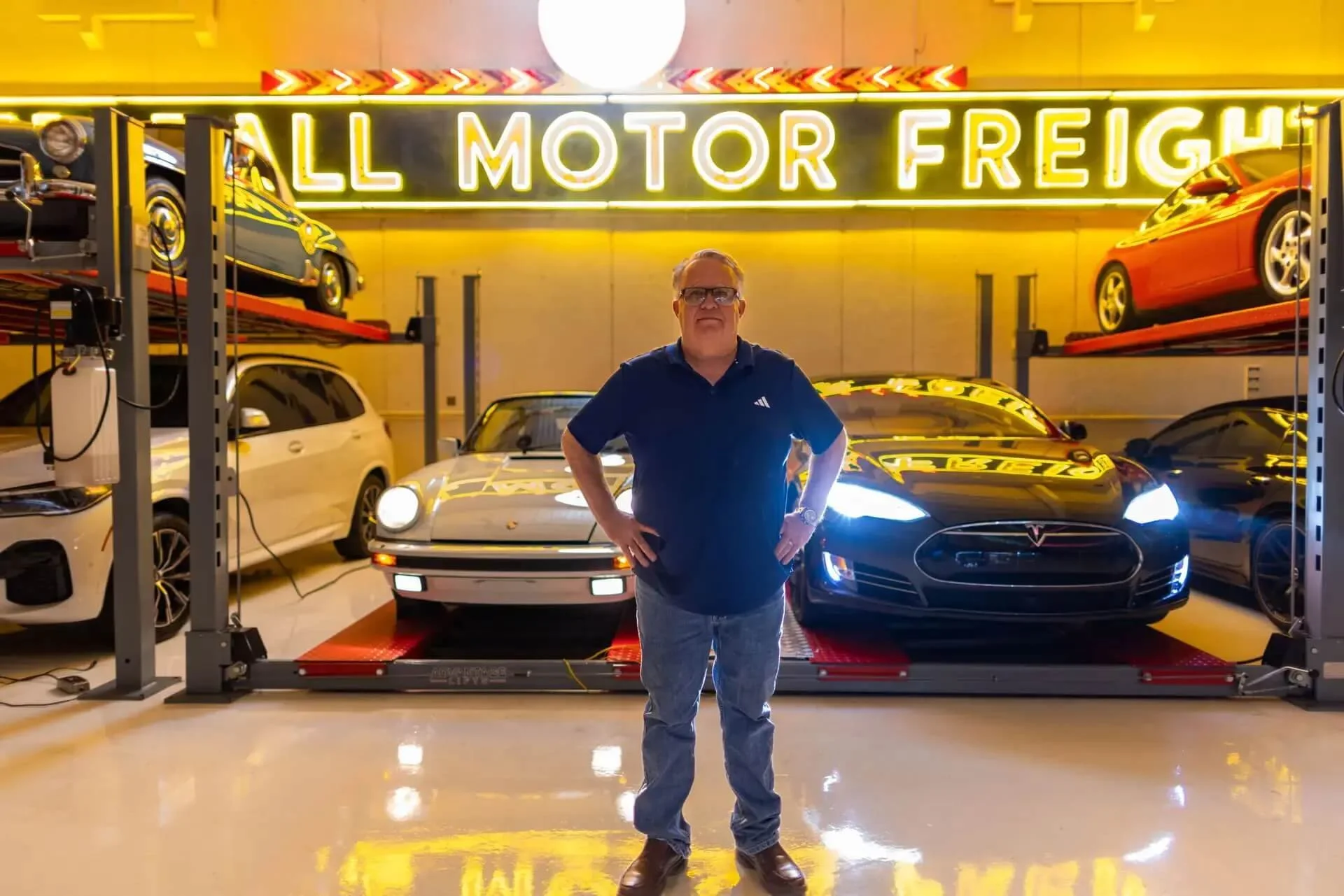 Man standing in front of cars in a showroom with yellow neon sign that reads 'AUTO MOTION MOTOR FREIGHT'.