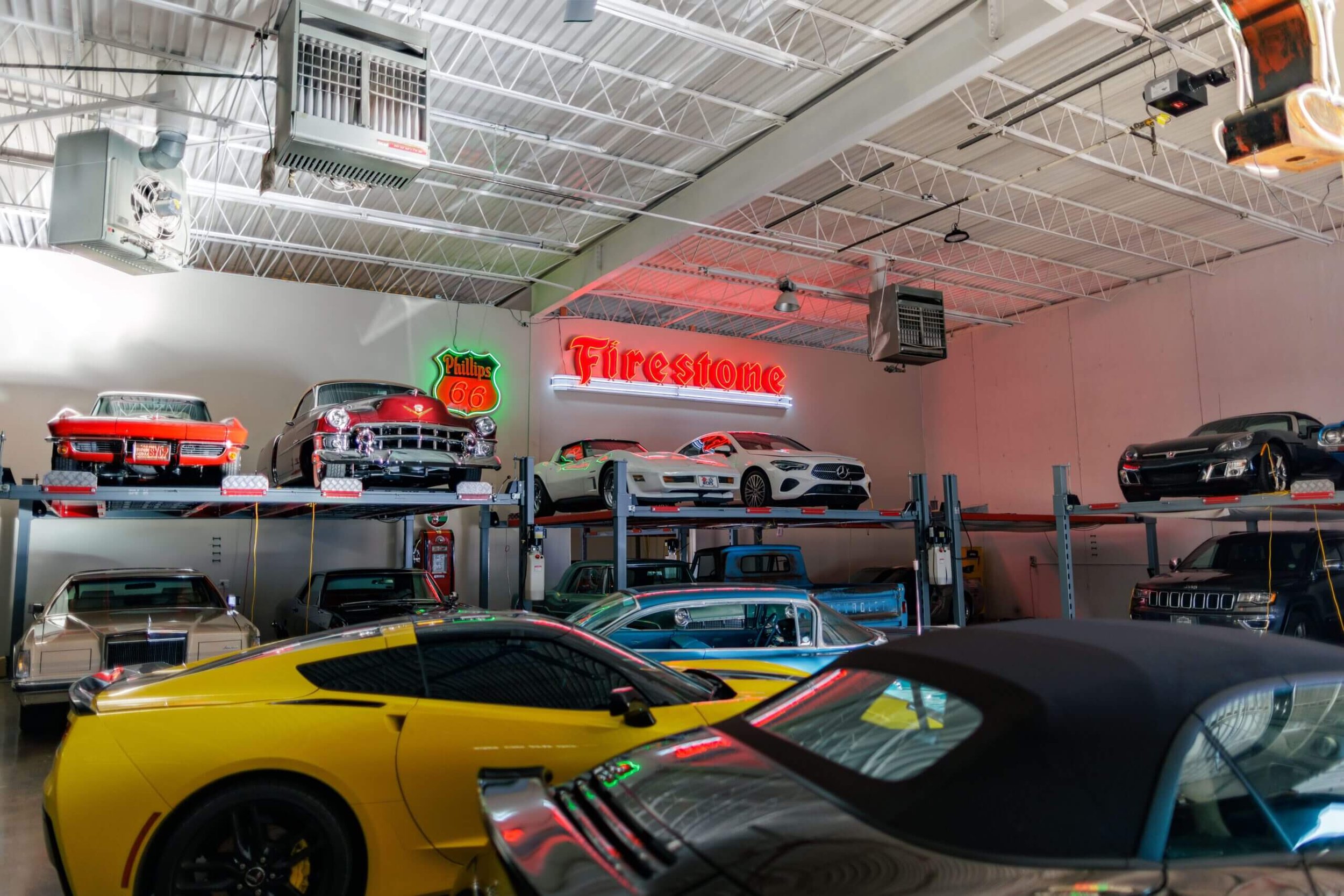 A collection of classic and modern cars in a garage or showroom with neon signs, including a Firestone sign and a Route 66 sign, and various car models displayed on elevated platforms.