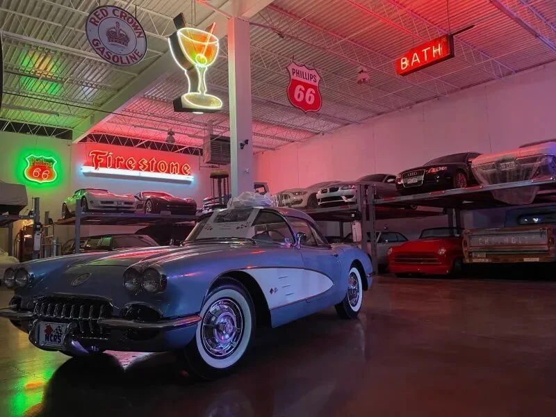 A vintage blue Corvette car in a garage decorated with neon signs and model cars on shelves, with signs reading 'Firestone,' 'Phillips 66,' and 'Bath'.