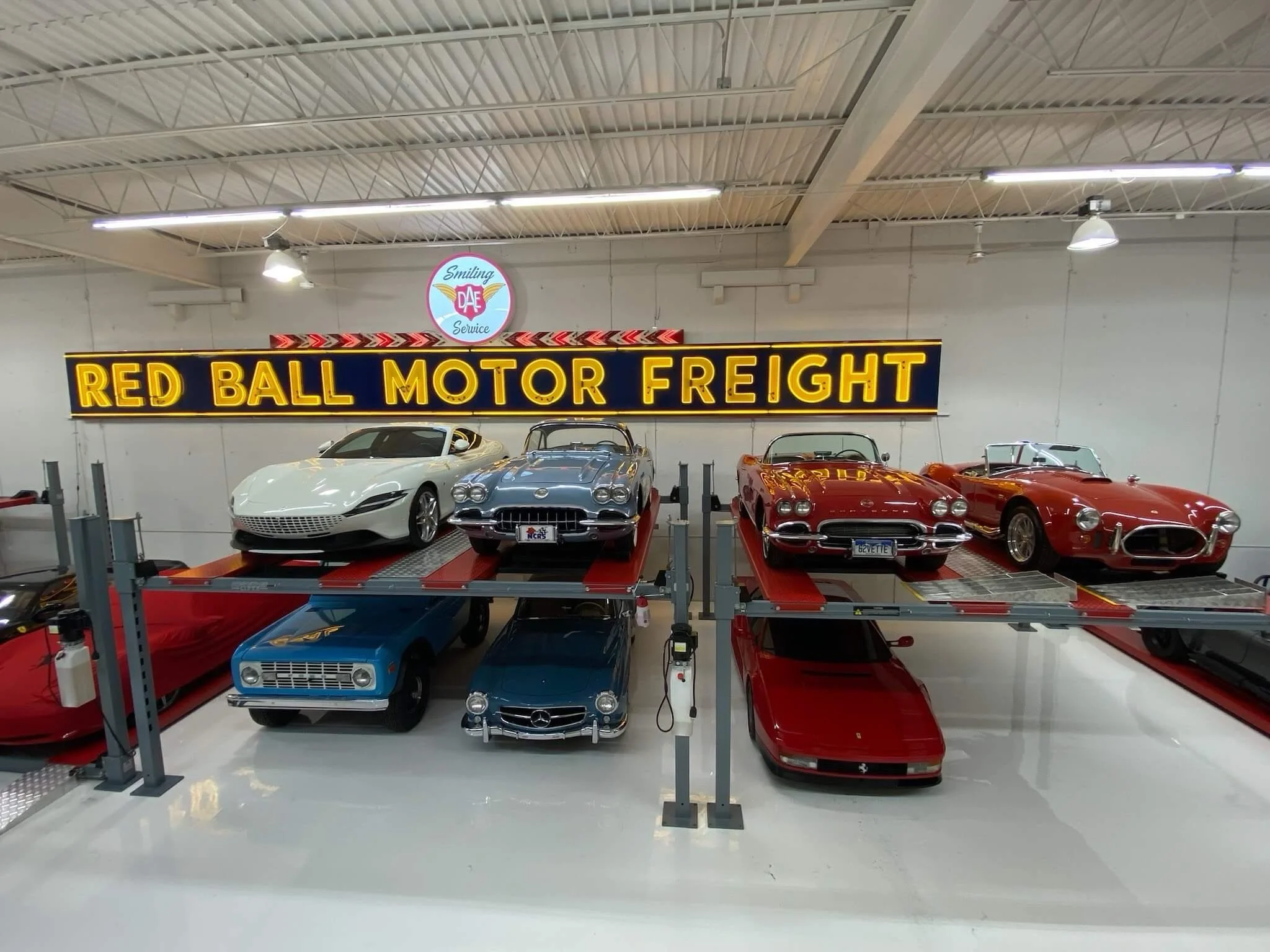 Collection of vintage and modern cars displayed in a warehouse with a sign reading 'Red Ball Motor Freight' and a circular sign that says 'Smiling DAE Service'.