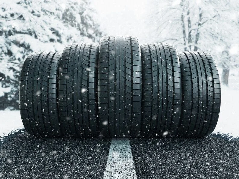 Six car tires lined up on a snow-covered road with fall trees in the background.