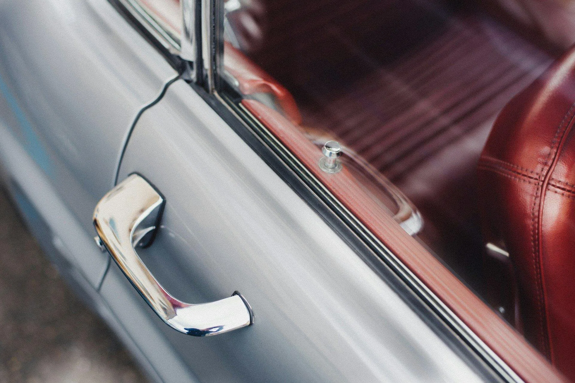 Close-up of part of a vintage car door with a chrome handle and a red leather interior.