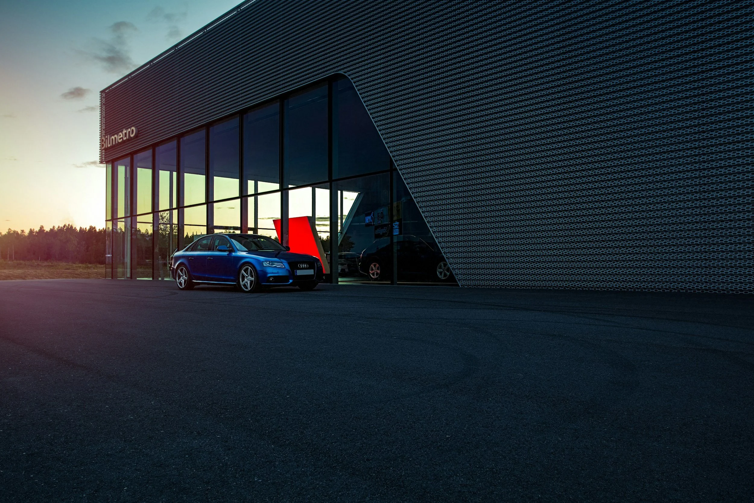 A blue car parked outside a modern glass building with the sign 'Bilmetro' at sunset.