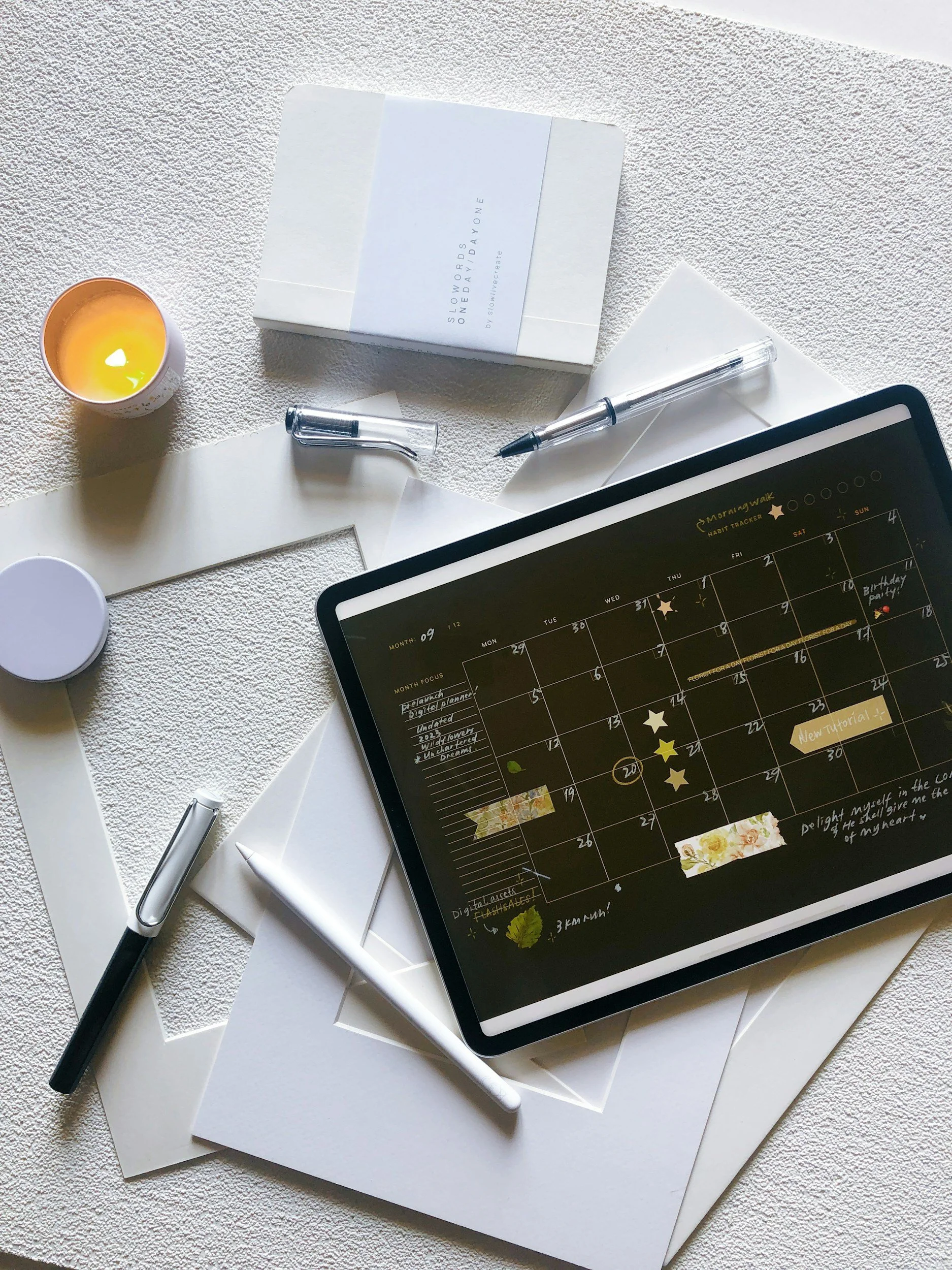 A flat lay of a workspace featuring a tablet displaying a digital calendar or planner, two pens, a white envelope, a white box labeled 'Slow Your Word One Day at One,' a candle, and some papers on a textured white surface.