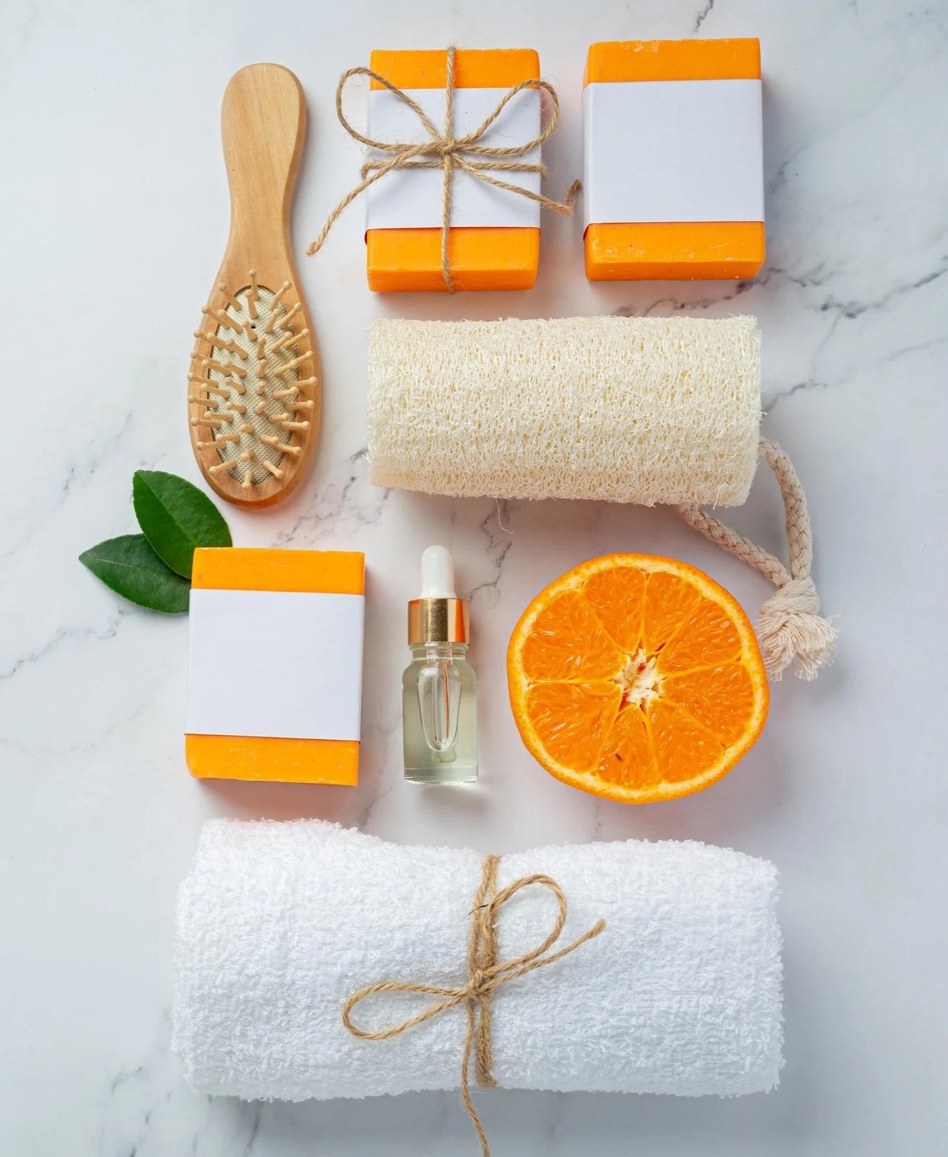 Arrangement of bath and skincare items, including soap bars, a loofah, a wooden brush, essential oil, a halved orange, and a rolled white towel on a marble surface.