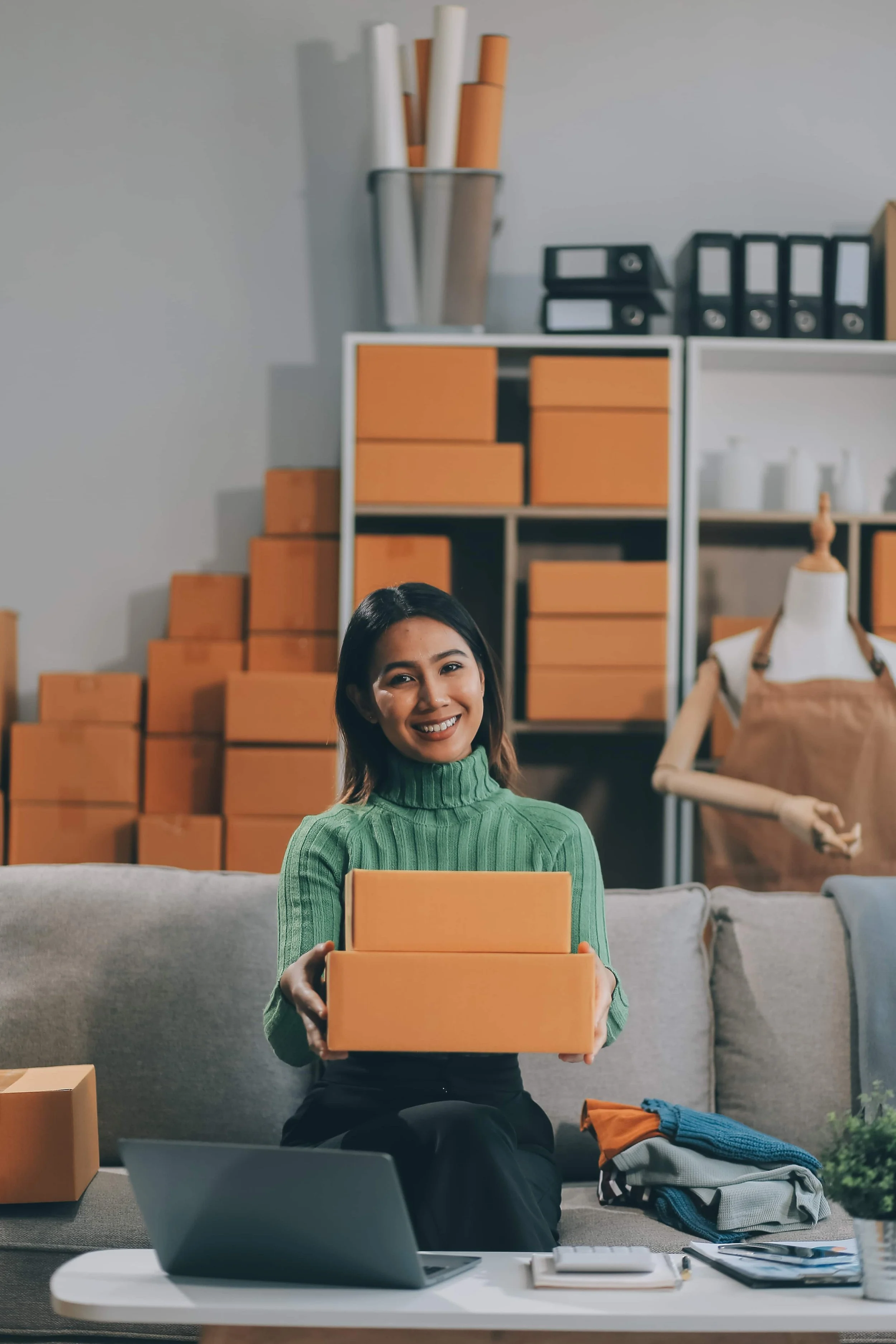 A woman sitting on a couch holding two orange boxes, with shelves of boxes and a mannequin in the background, in a workspace or storage area.