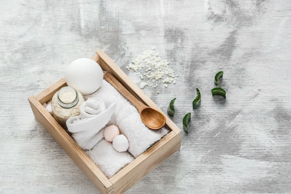 Spa tray with bath essentials including white eggs, wrapped towel, jar of bath salts, wooden spoon, green leaves, and white bath powder on a wooden surface.