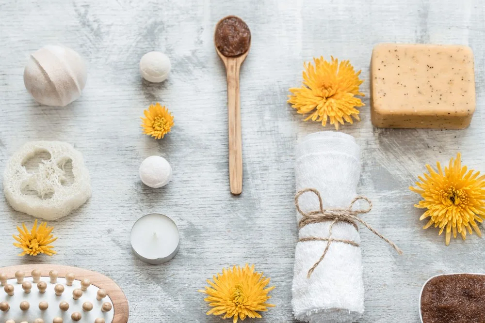Arrangement of spa and wellness items including bath bomb, foam sponge, small balls, candle, wooden spoon with scrub, essential oil soaps, and yellow flowers on a white wooden surface.