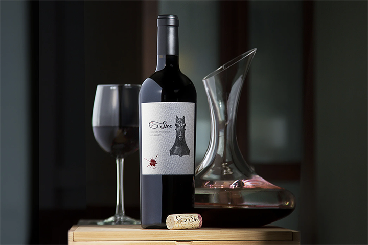 A bottle of red wine, a filled wine glass, a glass decanter, and a wine cork on a wooden surface.