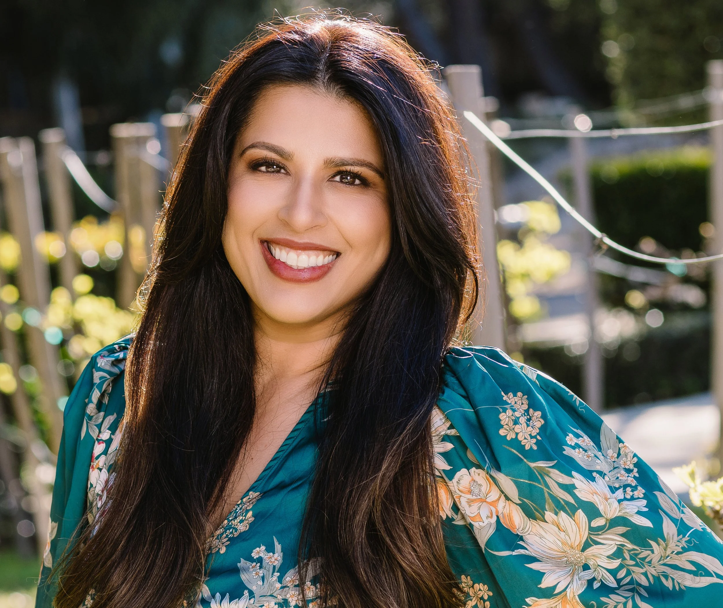 A woman with long dark hair and a bright smile, wearing a blue floral dress, outdoors in a sunny setting with blurred greenery in the background.